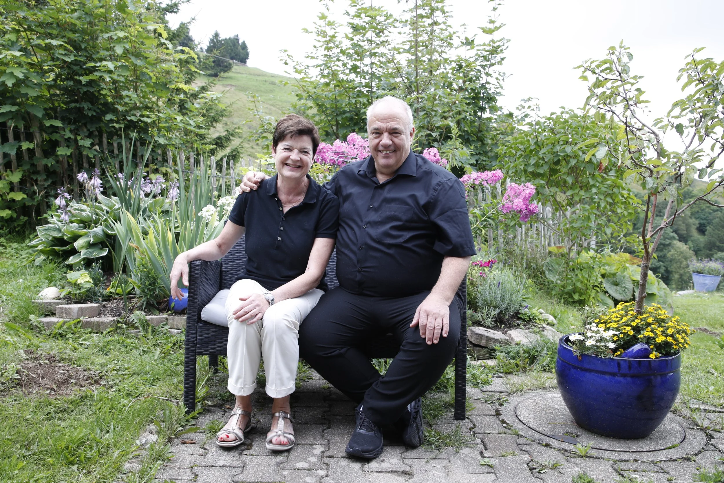 Ein lächelndes älteres Paar sitzt gemeinsam auf einer Bank im Garten, umgeben von Blumen und Büschen, mit grünem Hang im Hintergrund.