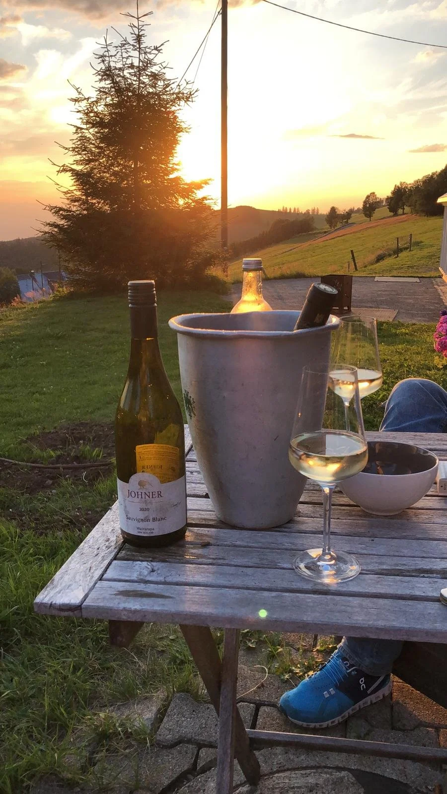 Sonnenuntergang, Weinflaschen, Weingläser mit Weißwein, Eimer mit Weinflasche, Holztablett, grüne Wiese, Baum, Hütte im Hintergrund.