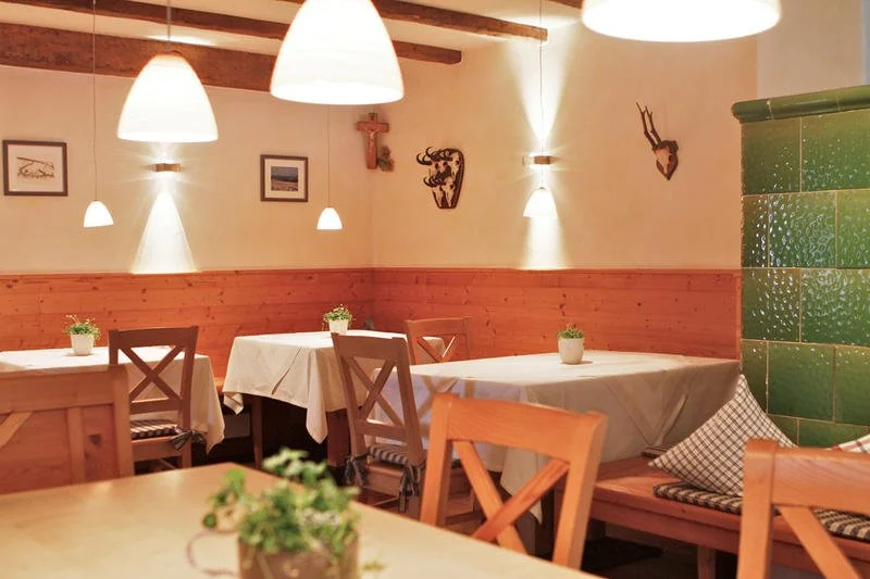 Ein gemütliches Restaurant mit Holzmöbeln, weißen Tischtüchern, kleinen Topfpflanzen auf den Tischen und warmem Licht. Wände mit Holzverkleidung und dekorativen Wandbildern sowie Wandbeesten. Ein grüner Ofen aus Kacheln auf der rechten Seite.