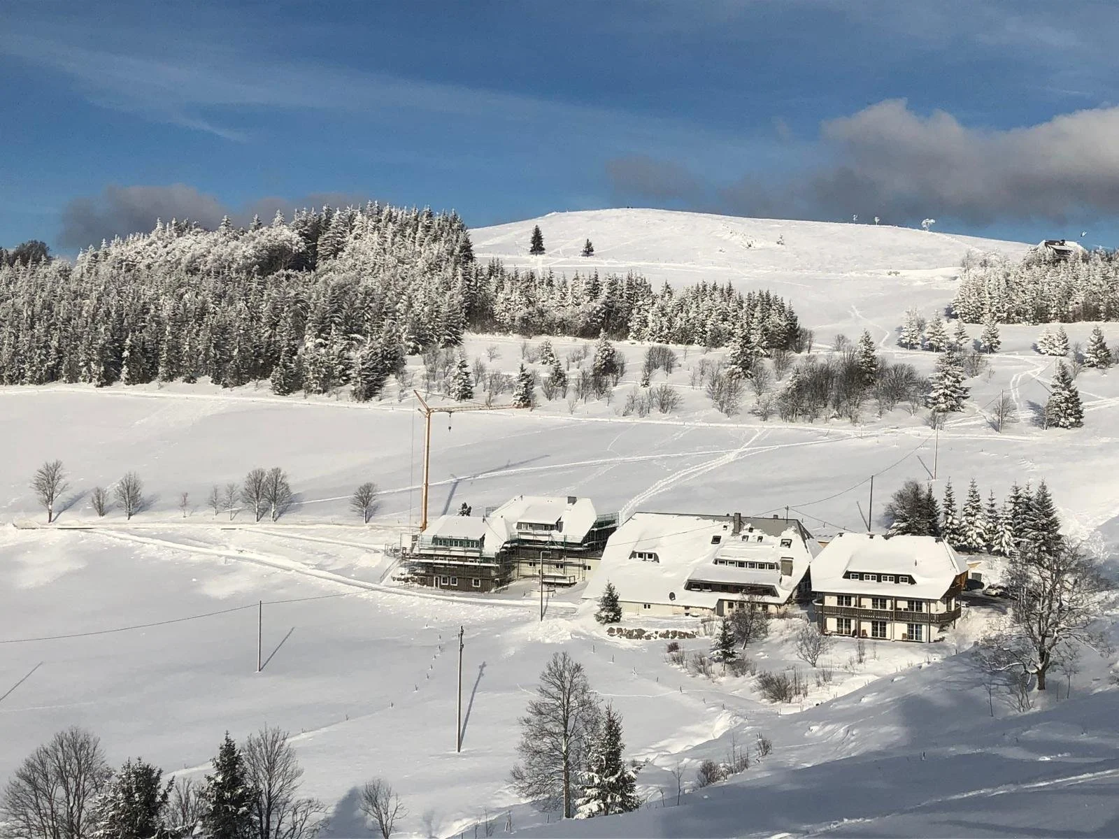 Schneebedeckte Hügellandschaft mit Häusern und einem Windrad, bewaldete Hügel im Hintergrund unter blauem Himmel.