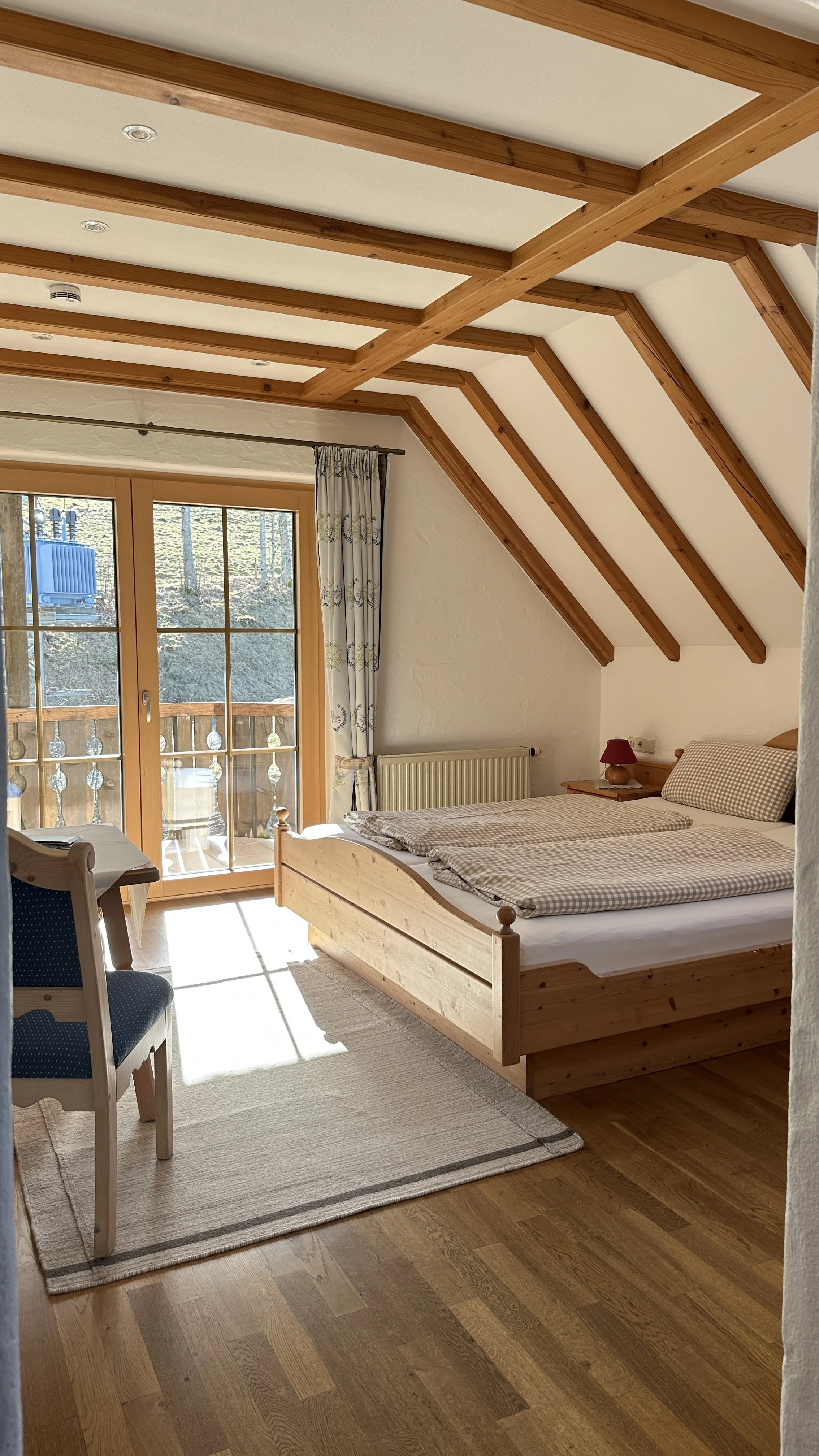 Ein Schlafzimmer mit Doppelbett, Holzbalken an der Decke, Balkon mit Holzgeländer, Fenster mit Blumenmuster-Vorhängen, kleiner Nachttisch mit rotem Lampenschirm, Sitzgelegenheit mit blauer Polsterung, Holzfußboden und helles Teppich