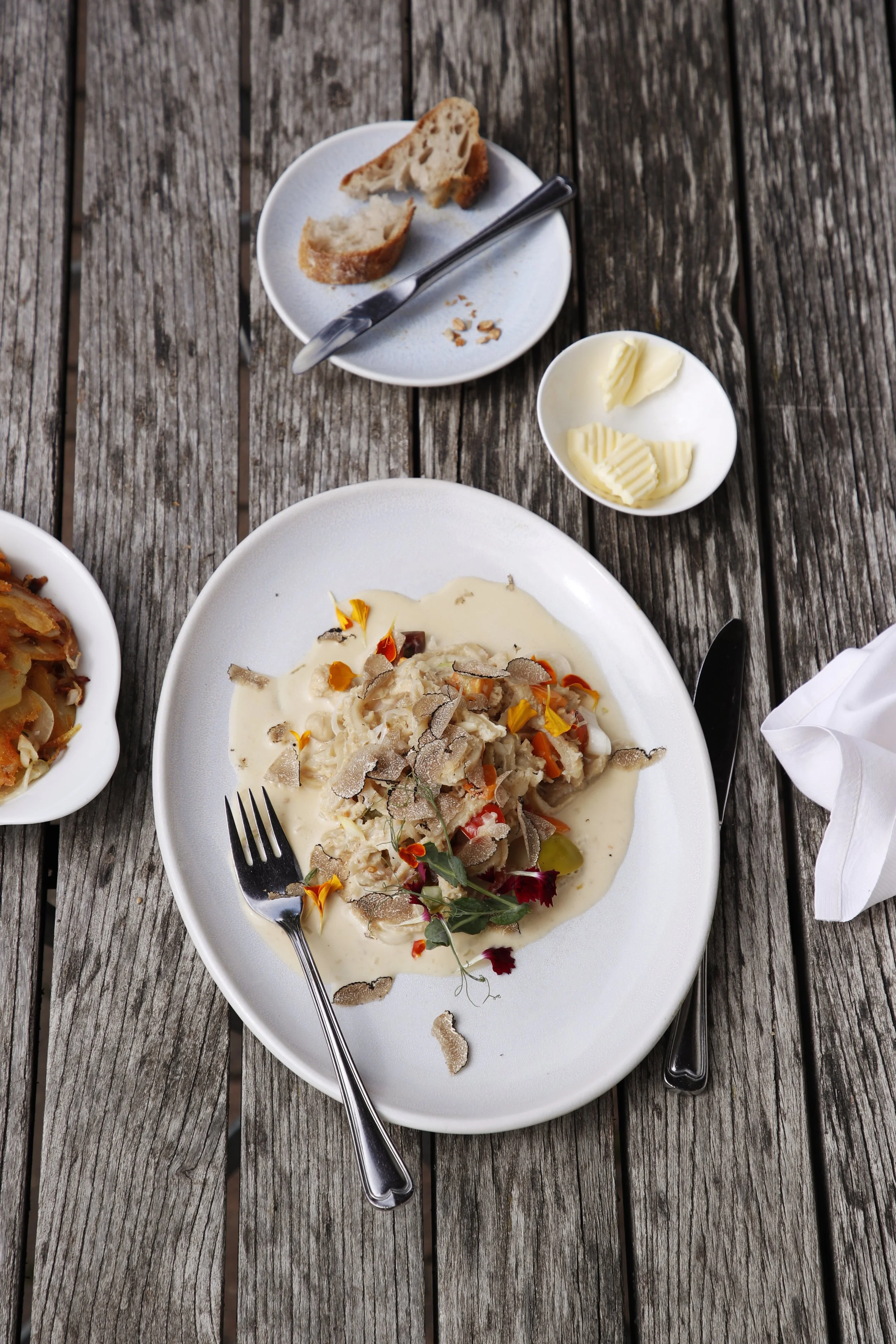 Ein Teller mit cremigem Gericht, garnieren mit Trüffelscheiben und Blättern, auf einem rustikalen Holztisch. Daneben Butter in einer kleinen Schüssel, und eine kleine Platte mit Brotscheiben, Messer und einige Brotsplitter.