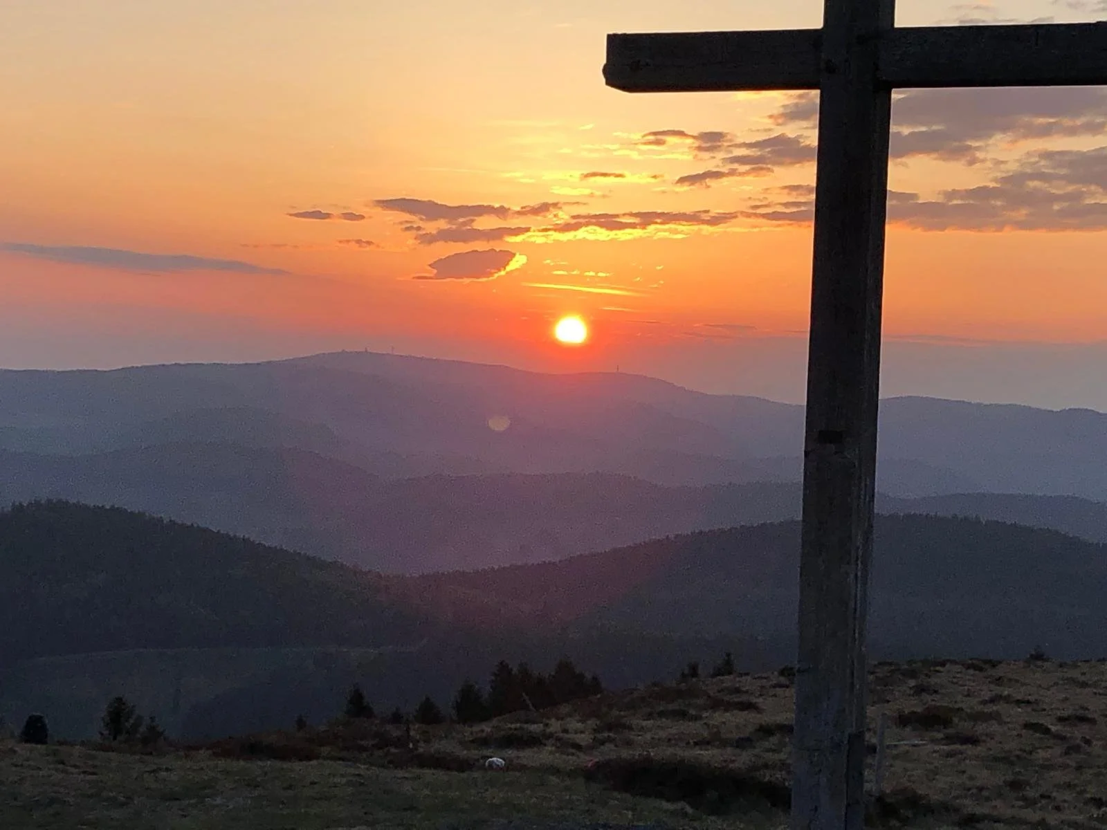 Berglandschaft bei Sonnenuntergang mit Himmel in warmen Farben und Schatten auf den Bergen, sichtbarer Holzkreuz im Vordergrund.