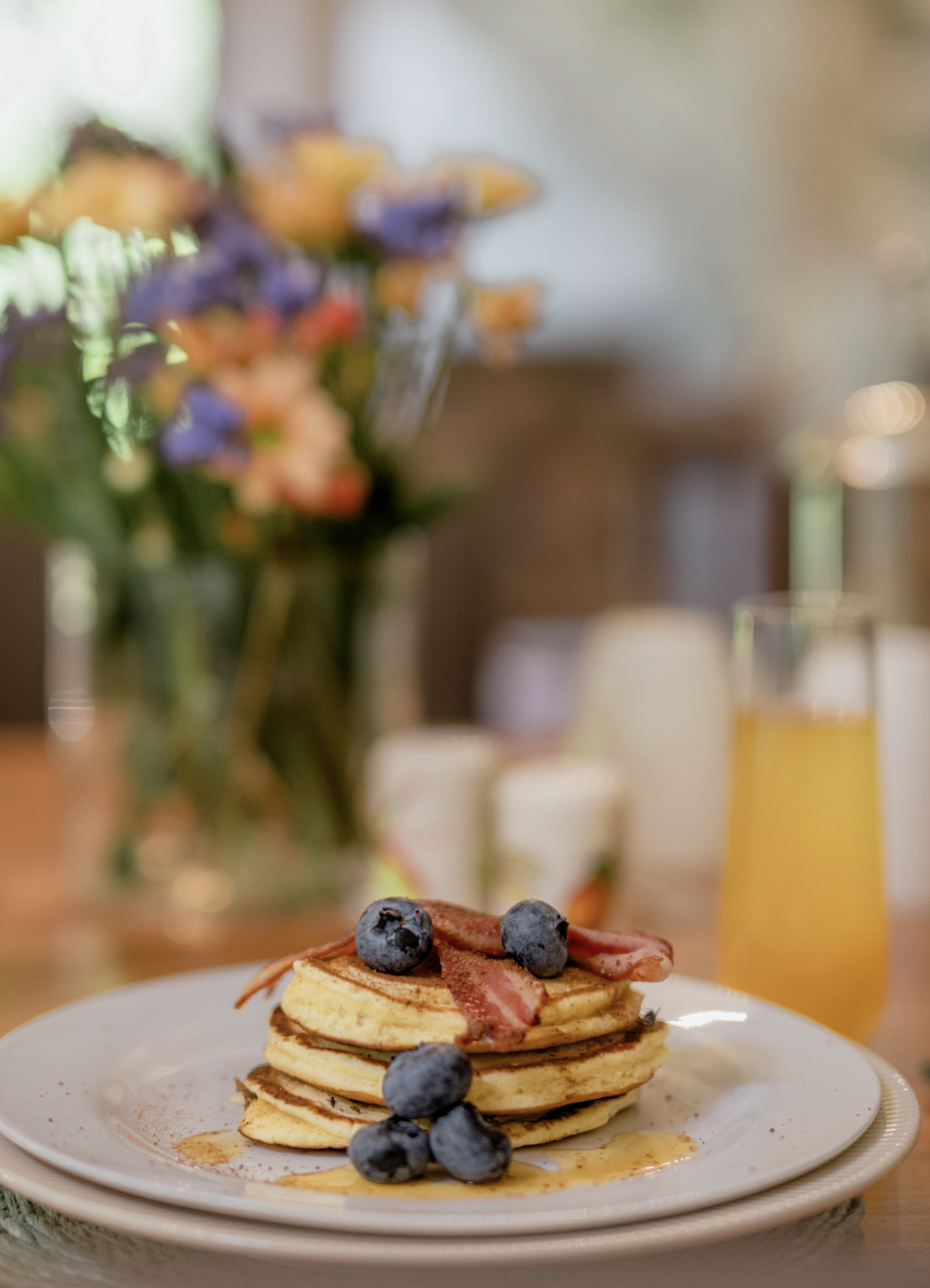 A stack of four pancakes topped with blueberries, bacon strips, and syrup on a white plate, with a blurred background of a floral arrangement and drinks.