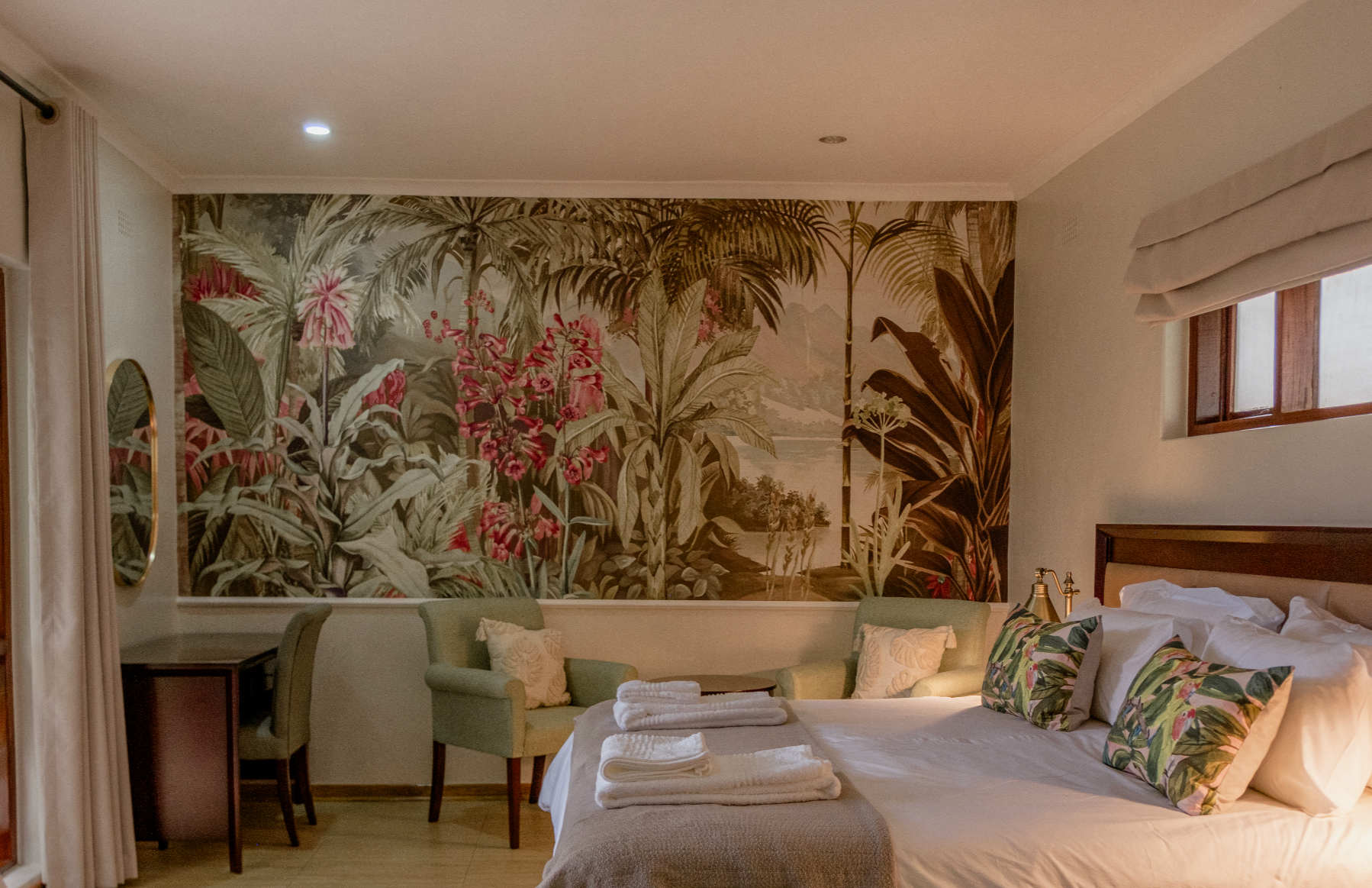 A bedroom with a large mural of a tropical jungle scene and pink flowers on the wall behind a bed with floral pillows. There are two green armchairs with pillows, a small wooden desk, and a window with beige Roman shades.