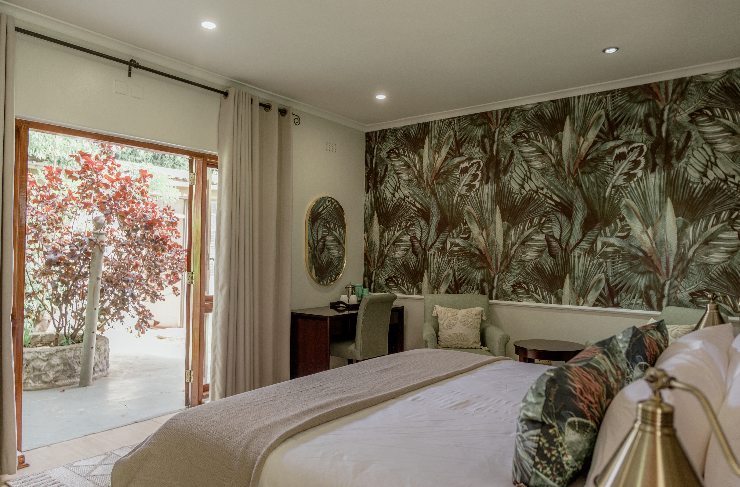 A cozy hotel bedroom with a bed, armchair, small desk, and a balcony door with curtains, overlooking a garden with a red-leafed tree.