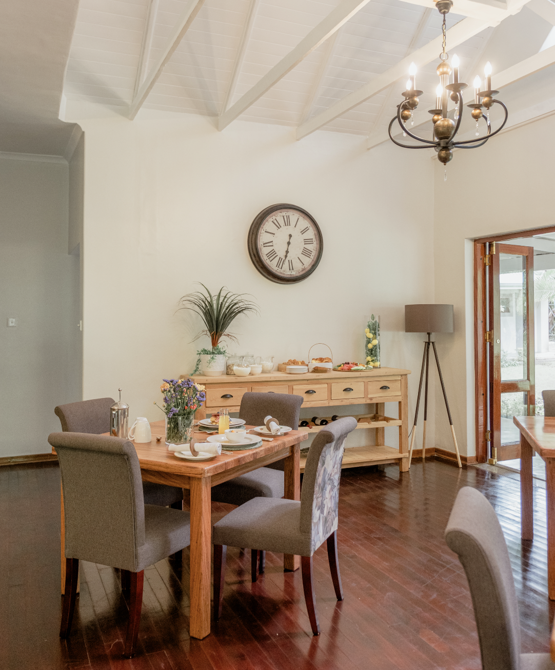 A dining room setup with a wooden table, gray upholstered chairs, a sideboard with food and drinks, a large clock on the wall, a chandelier overhead, and a door leading outside.