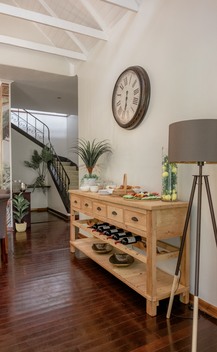 A wooden sideboard with wine bottles, bowls, and snack plates, near a wall with a large round clock. Potted plants and a floor lamp are also visible inside a house with hardwood flooring.