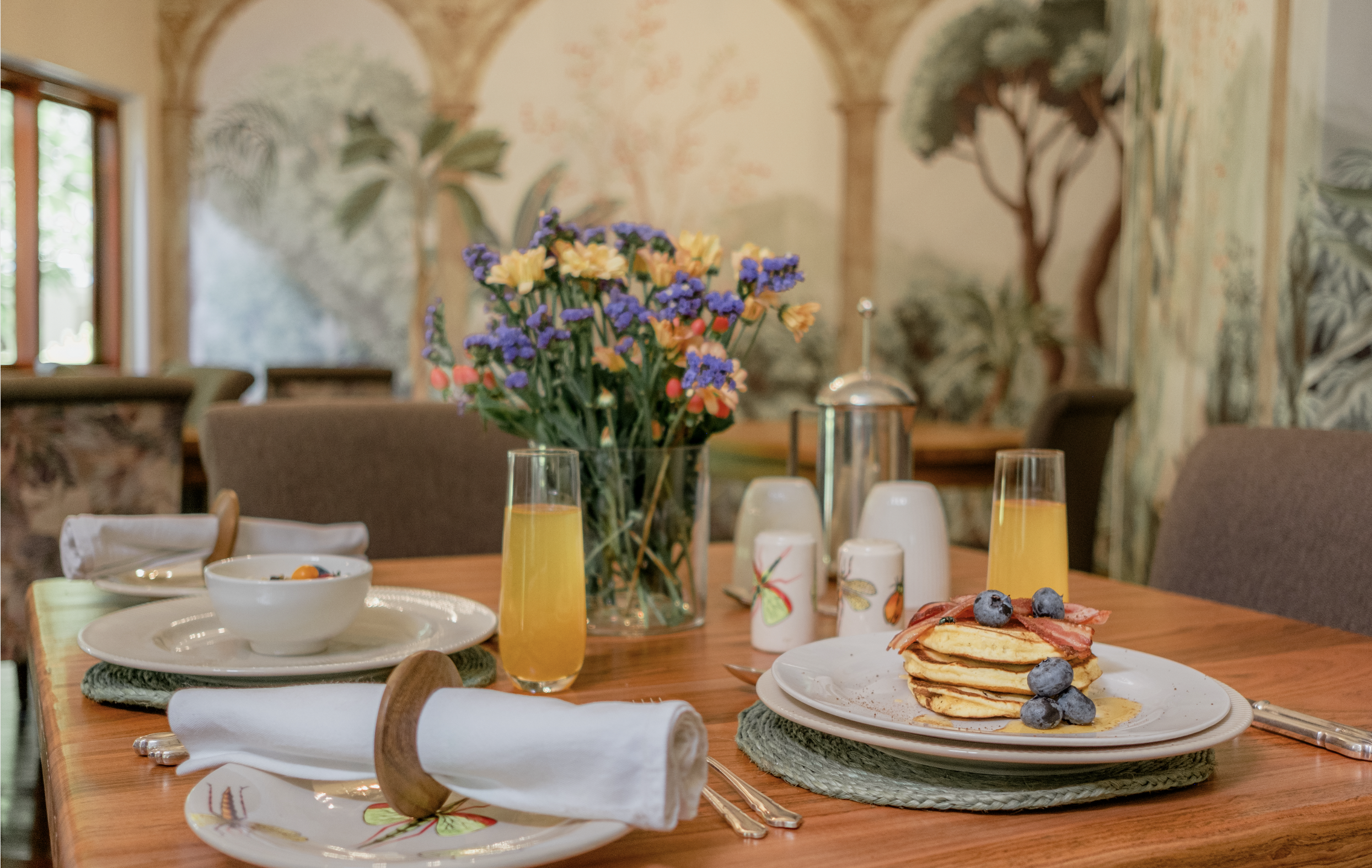A breakfast table set with a stack of pancakes topped with blueberries and bacon, two glasses of orange juice, a vase of colorful flowers, and various breakfast condiments, in a decorated dining room.