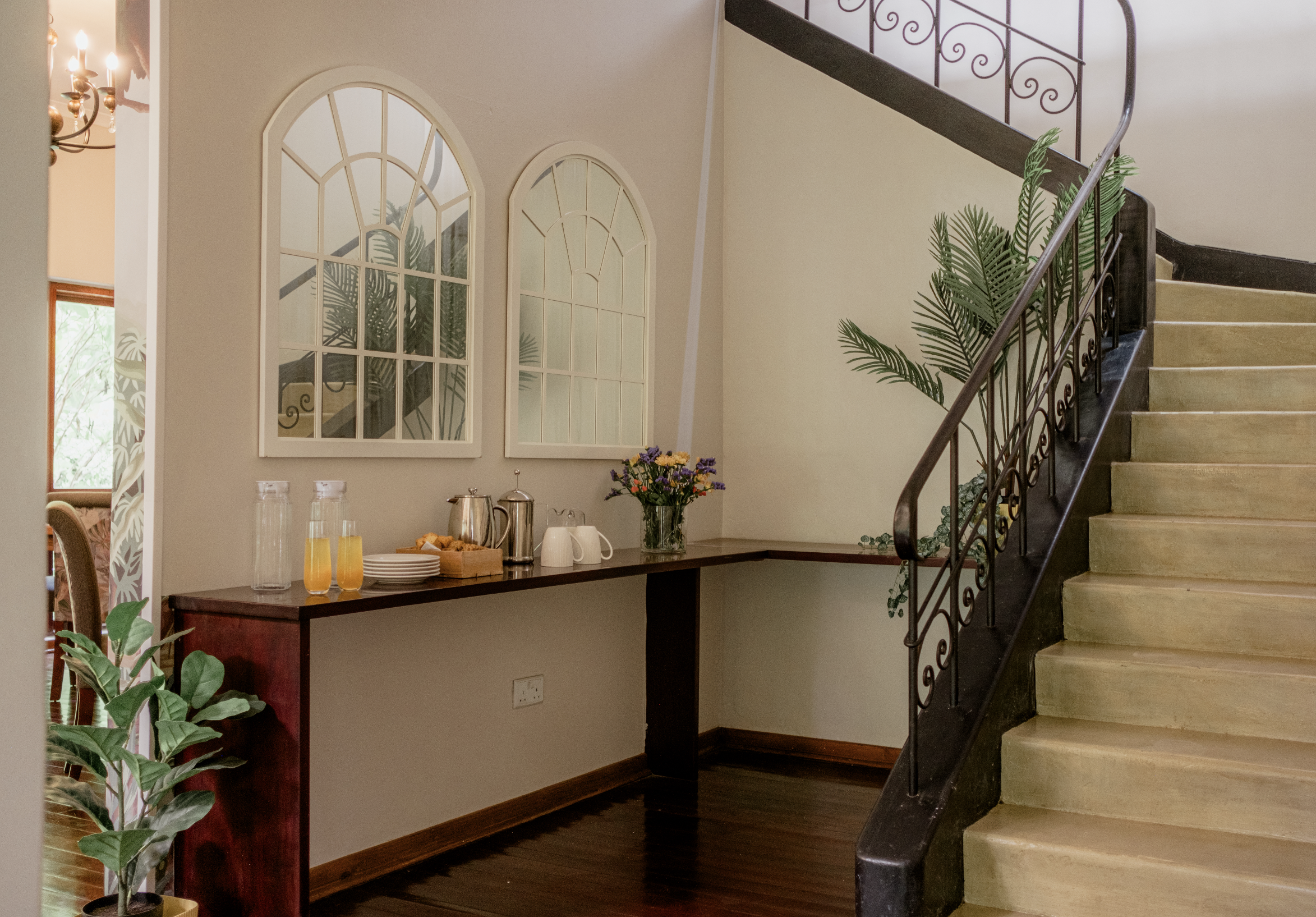 A staircase with black ornate railing leading up, a wooden floor, a wall with two arched windows reflecting a plant, a sideboard with a flower vase, juice bottles, plates, cups, and a coffee pot.