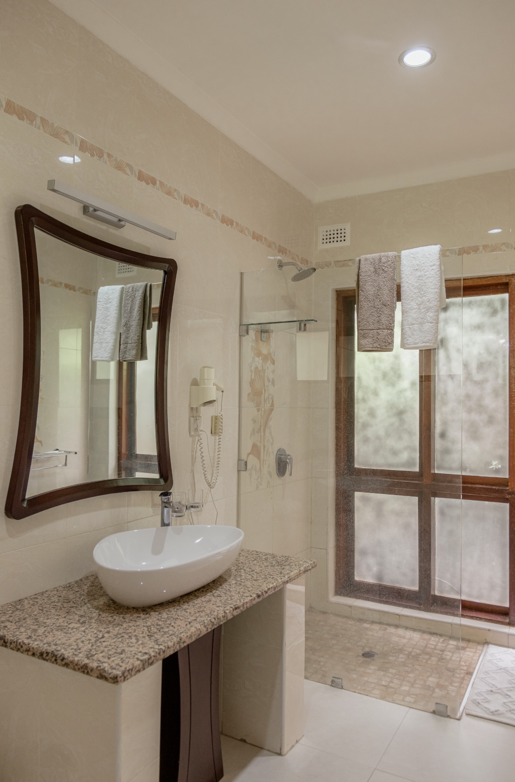 A bathroom with a wavy mirror, a vessel sink on a granite countertop, a glass-enclosed shower area with towel bars, a window with wooden framing, and a wall-mounted hair dryer.