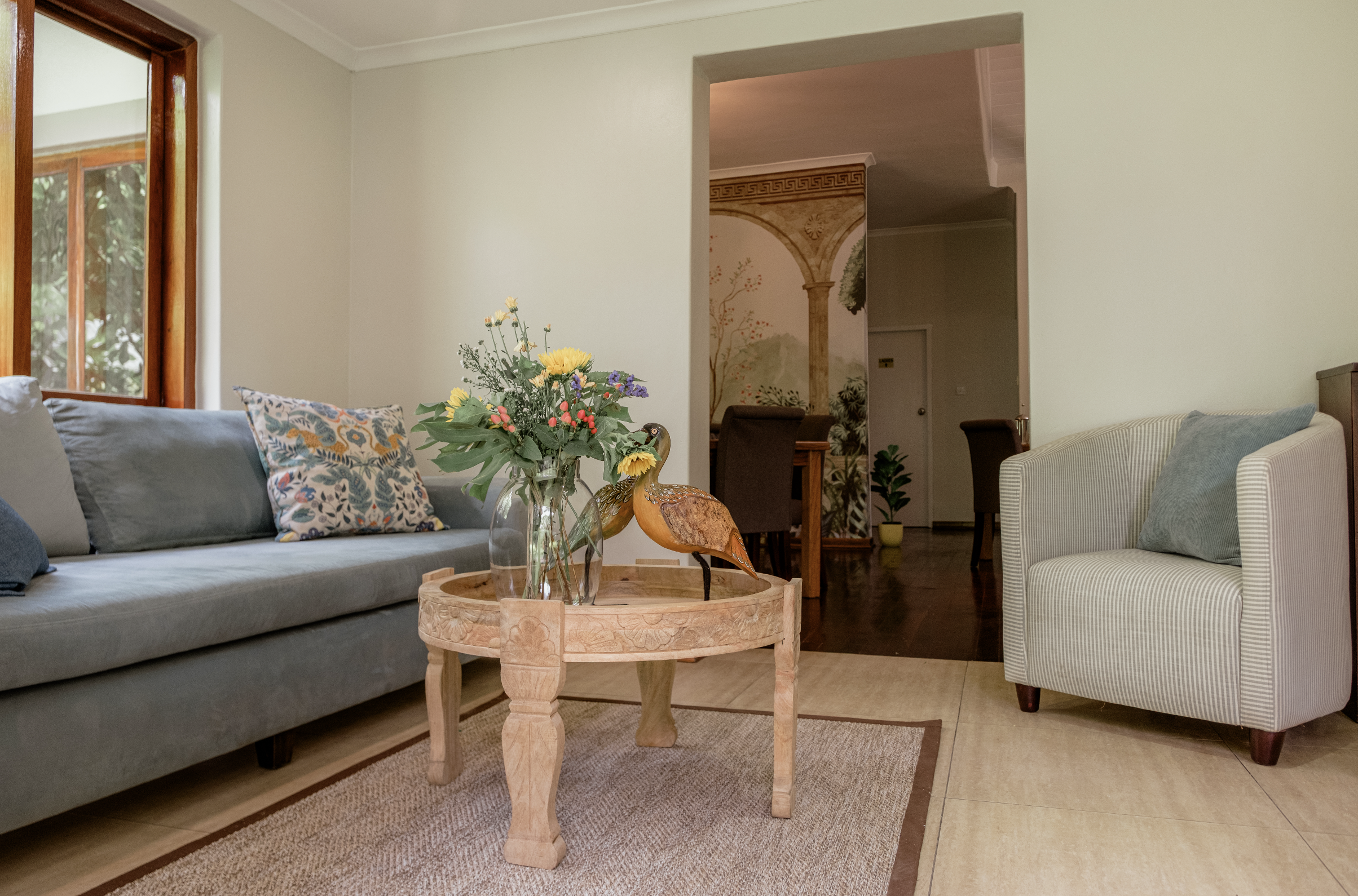 Living room with gray sofa, patterned cushion, striped armchair, wooden coffee table with glass vase of flowers and decorative ducks, window with wooden frame, and view into dining area with mural depicting a classical scene.