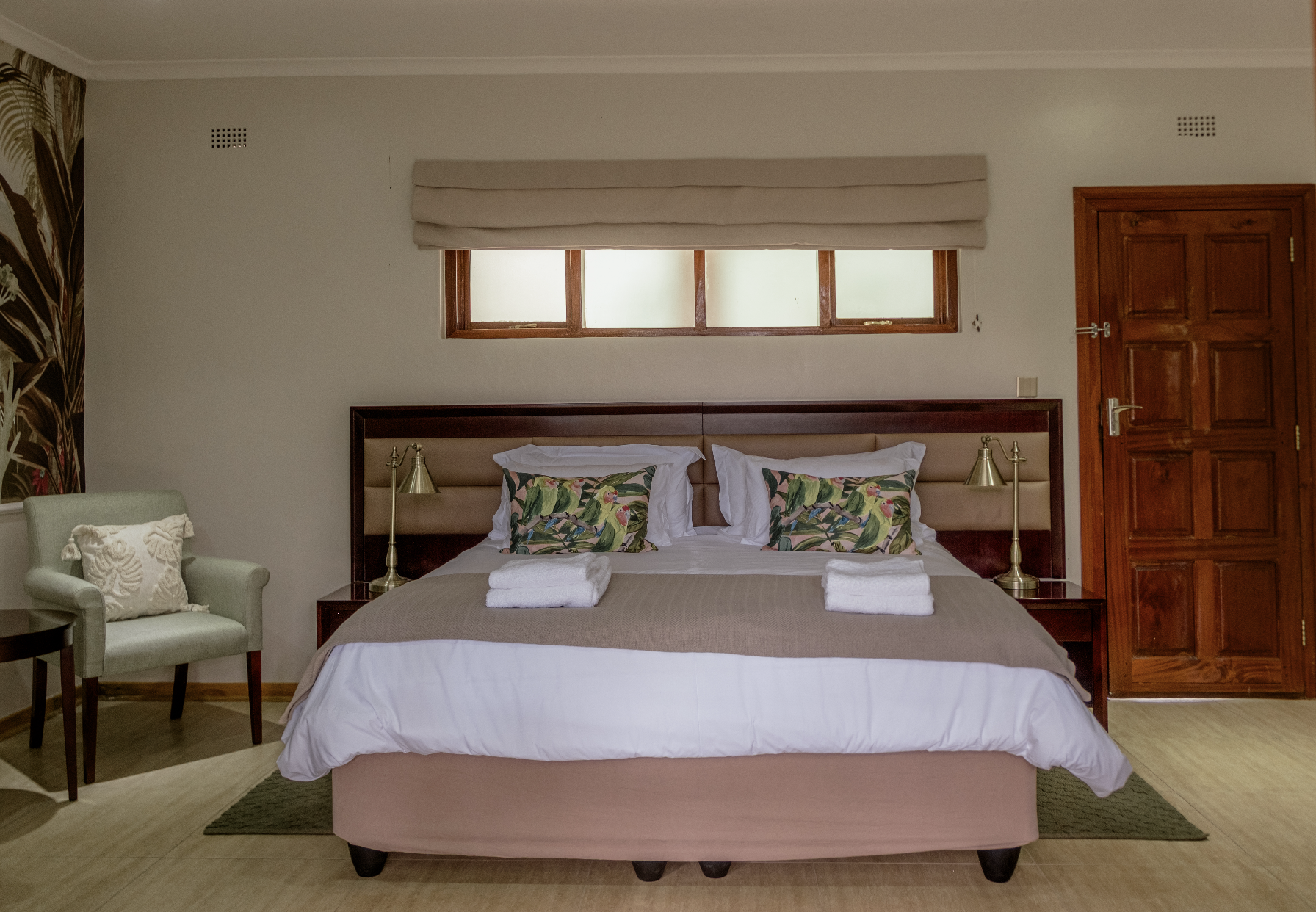 Hotel room with a large bed, decorative pillows with tropical pattern, wooden door, window above the bed, beige walls, armchair with pillow, and bedside lamps.