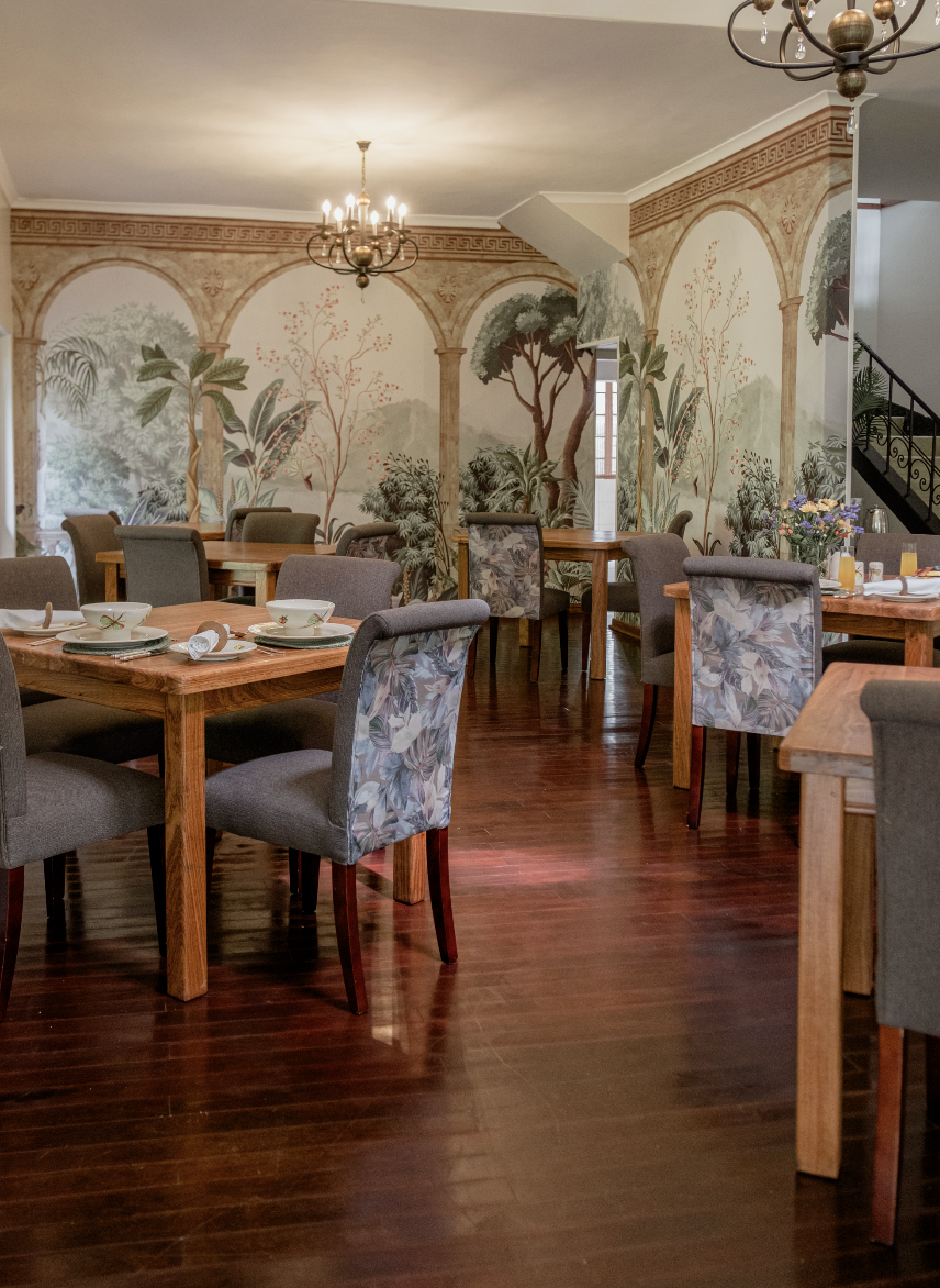 Elegant dining room with wooden tables, upholstered chairs, decorative wall mural of nature scenes, and chandeliers hanging from the ceiling.