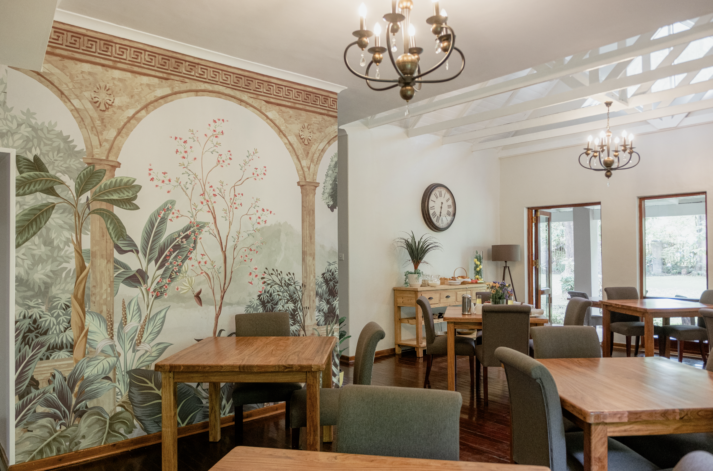 Interior of a dining area with wooden tables and chairs, decorative wall mural featuring trees and plants, ceiling chandeliers, a wall clock, and a door leading outside.