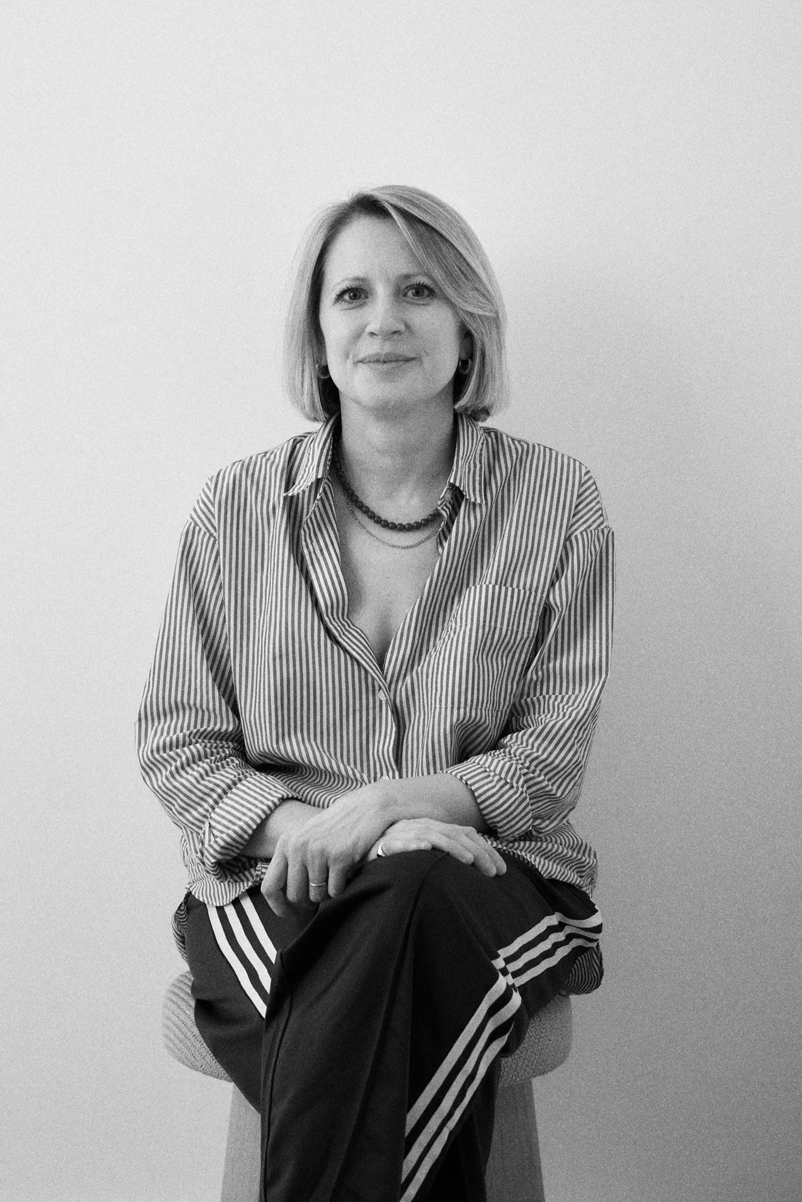 A woman sitting on a chair against a plain wall, wearing a striped shirt, jeans, and a necklace, with her hands resting on her lap.