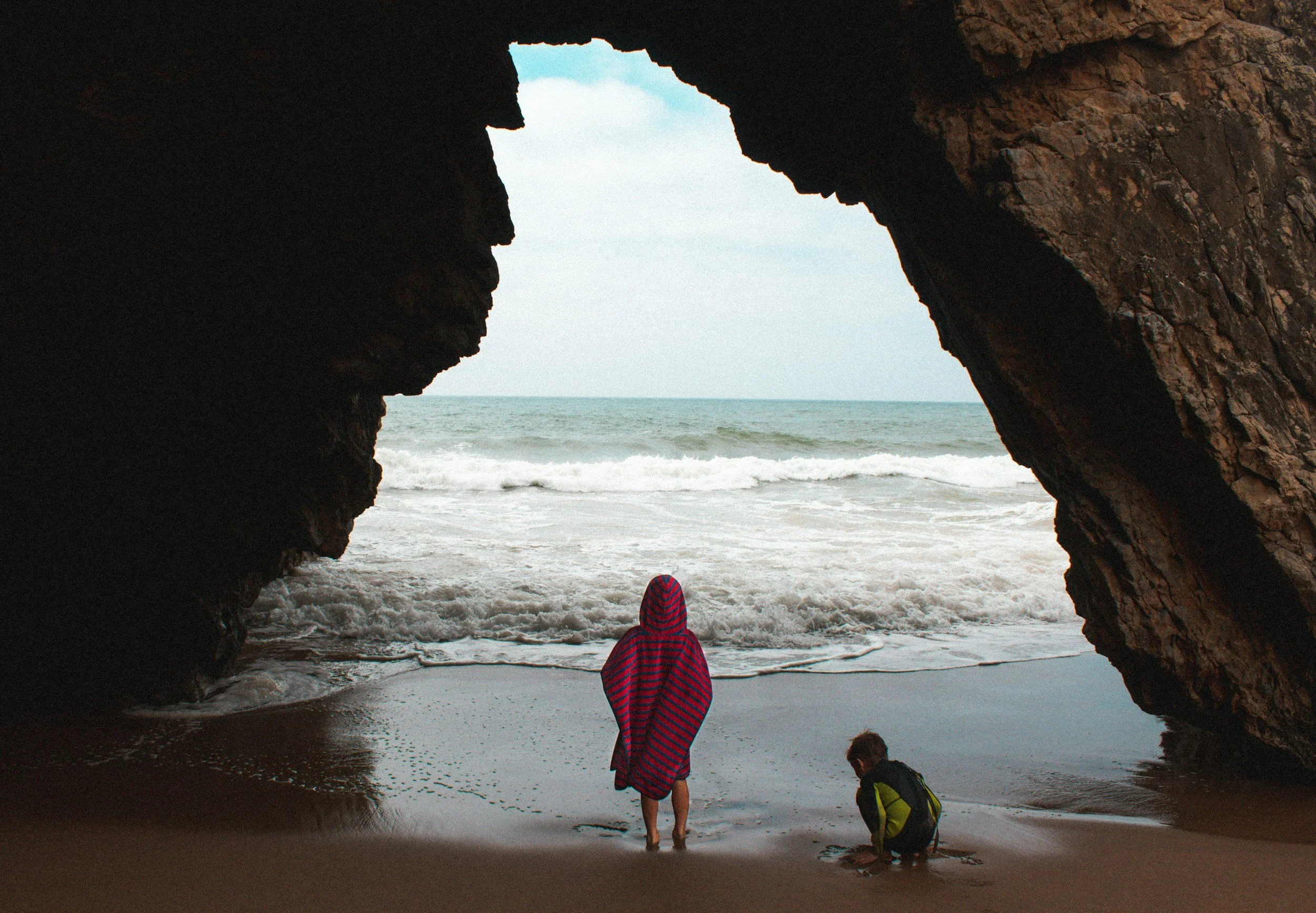 View from inside a rocky cave looking out onto the ocean, with three children playing on the beach near the water.