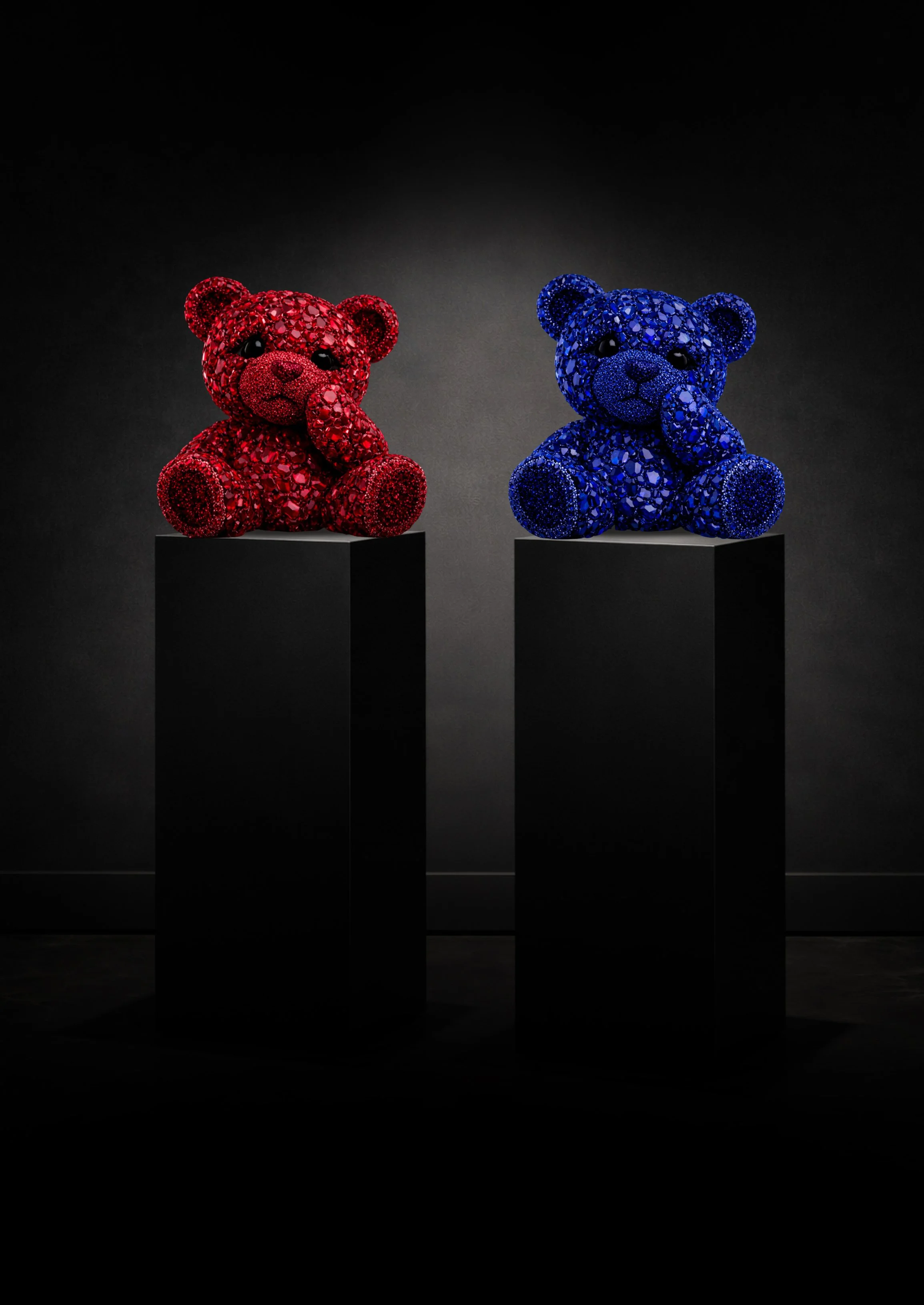Two teddy bear sculptures, one red and one blue, covered in shiny gemstones, sitting on black pedestals against a dark background.