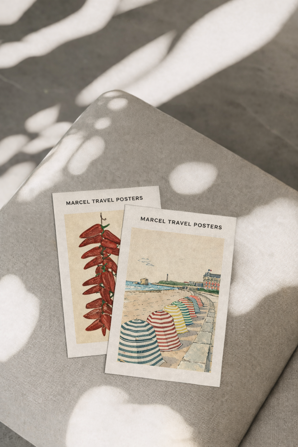 Deux affiches de voyage de Marcel, une avec des piments rouges et l'autre avec des parasols colorés sur une plage, posées sur une surface gris clair avec des taches de lumière