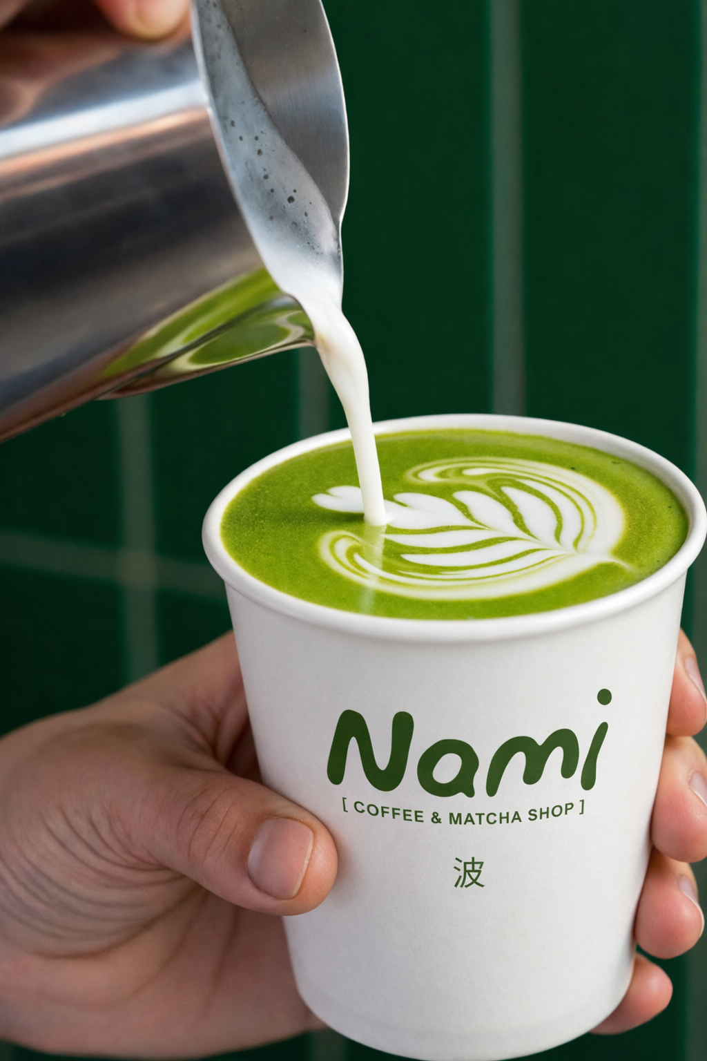 Une main tient un gobelet en papier avec la marque 'Nami' et la description '[ Coffee & Matcha Shop ]'. Un liquide vert est versé dans le gobelet, créant un motif artistique de latte sur la surface, probablement un matcha latte.