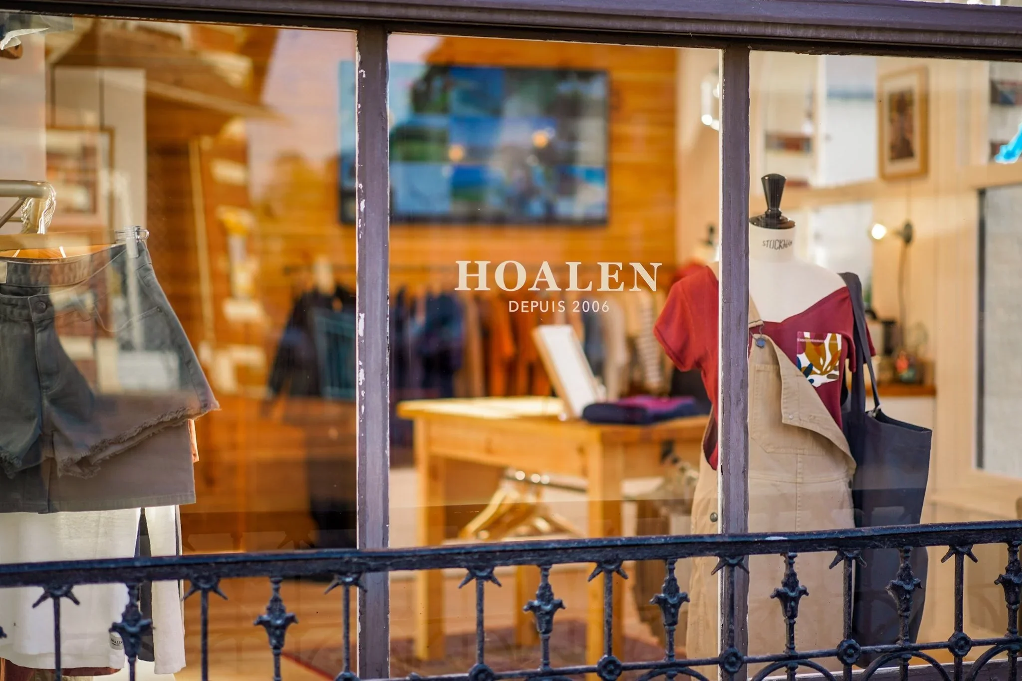 Vitrine d'une boutique de vêtements avec mannequins portant des vêtements à l'extérieur, en bois et métal, avec le nom 'HOALEN' et 'DEPUIS 2006' écrit sur la vitre.