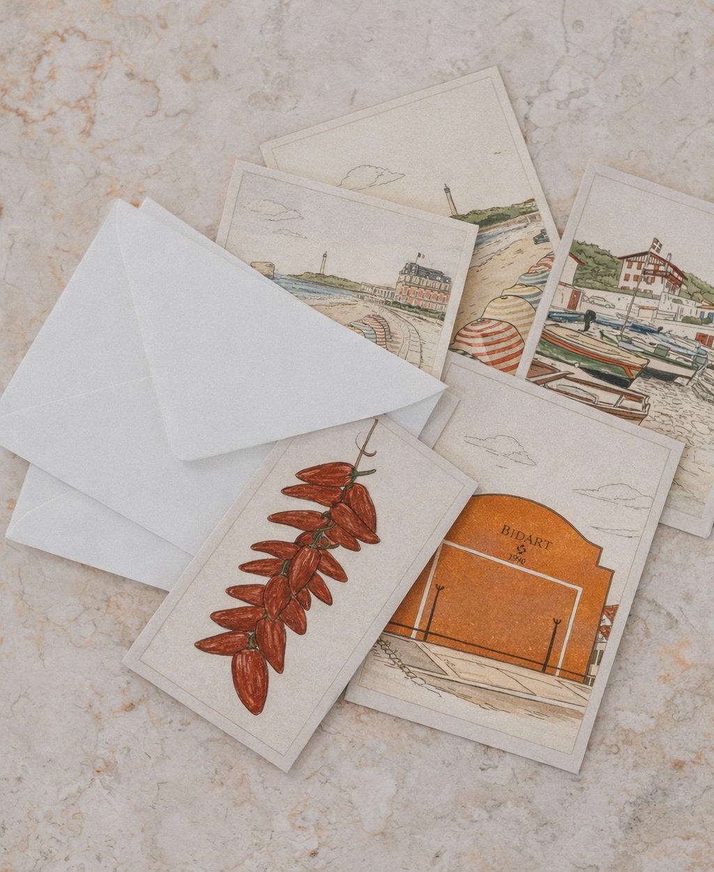 Cartes postales illustrées représentant des paysages côtiers avec des bateaux, un bâton de cayenne séché, et une enveloppe blanche.