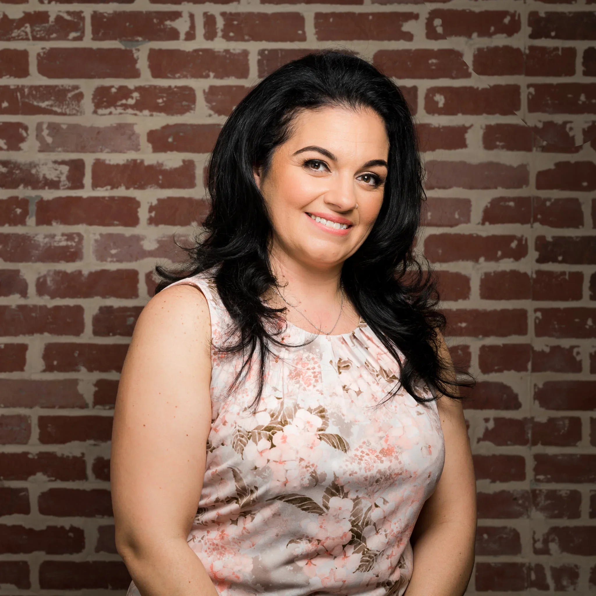 A woman with long black hair, wearing a sleeveless floral top, smiling, standing in front of a brick wall background.