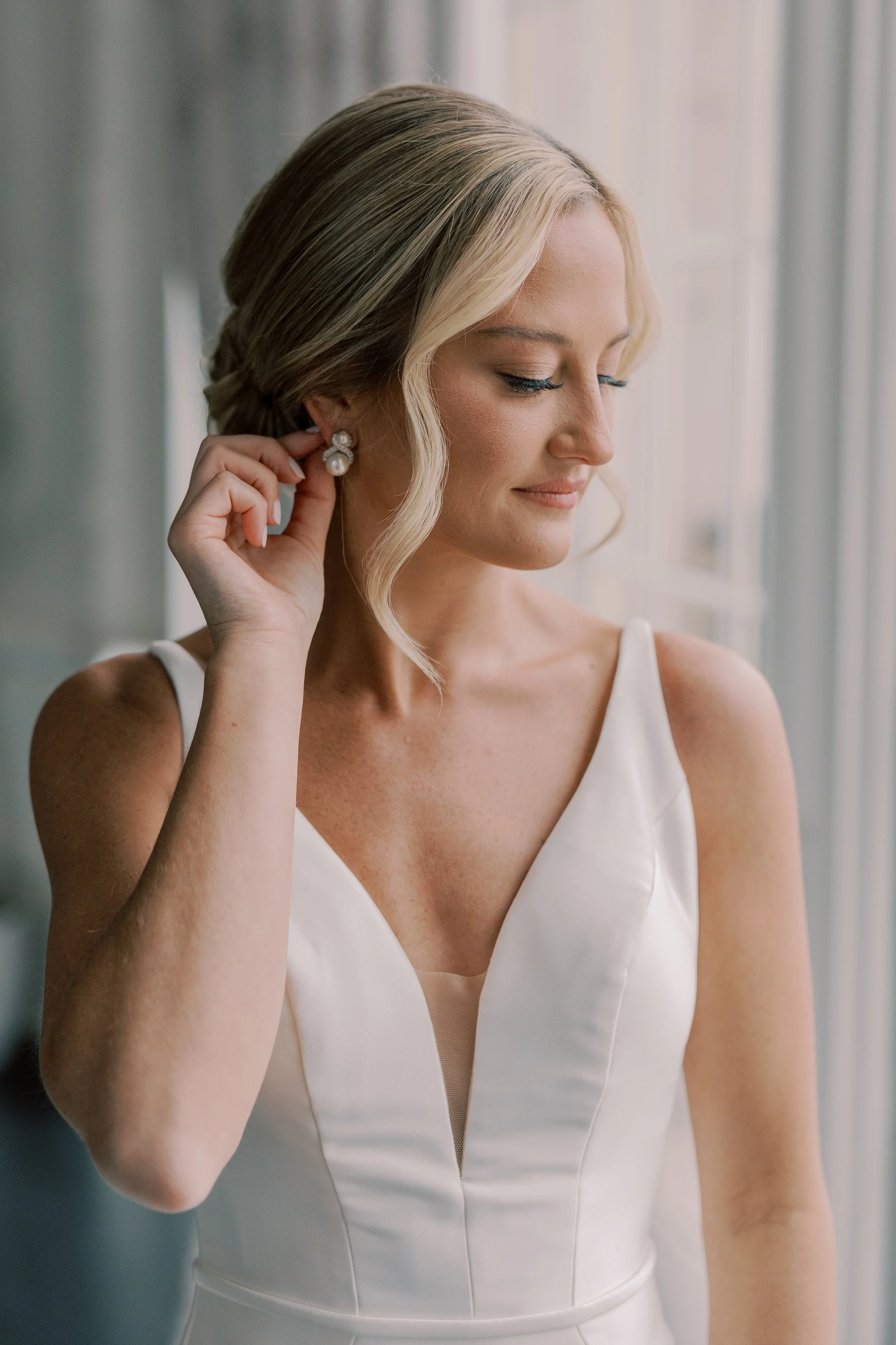 A woman in a white dressPutting on earrings by a window.