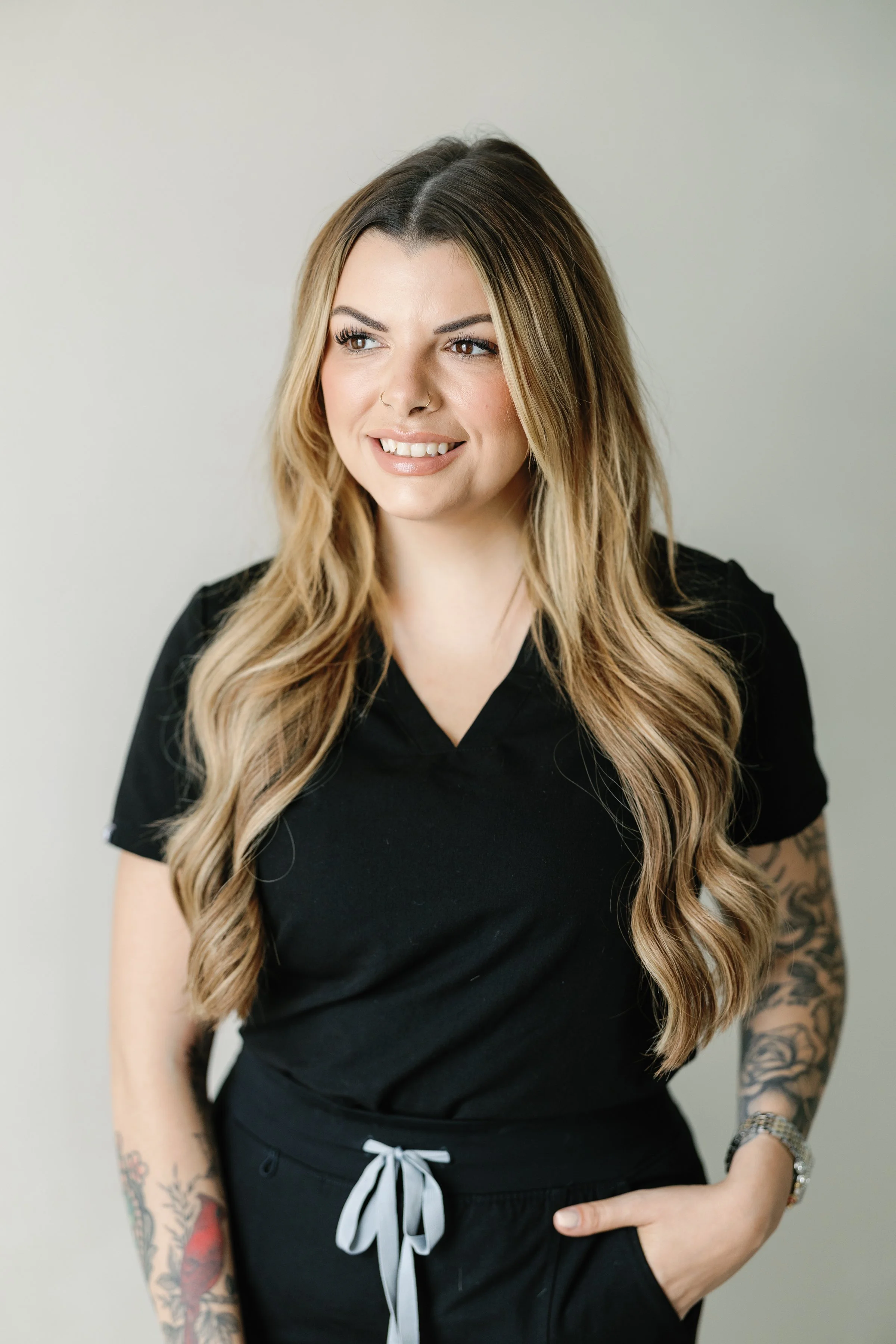 A woman with long, wavy, blonde hair wearing a black medical scrub top, with tattoos on her right arm and a watch on her left wrist, smiling and looking slightly to the side.