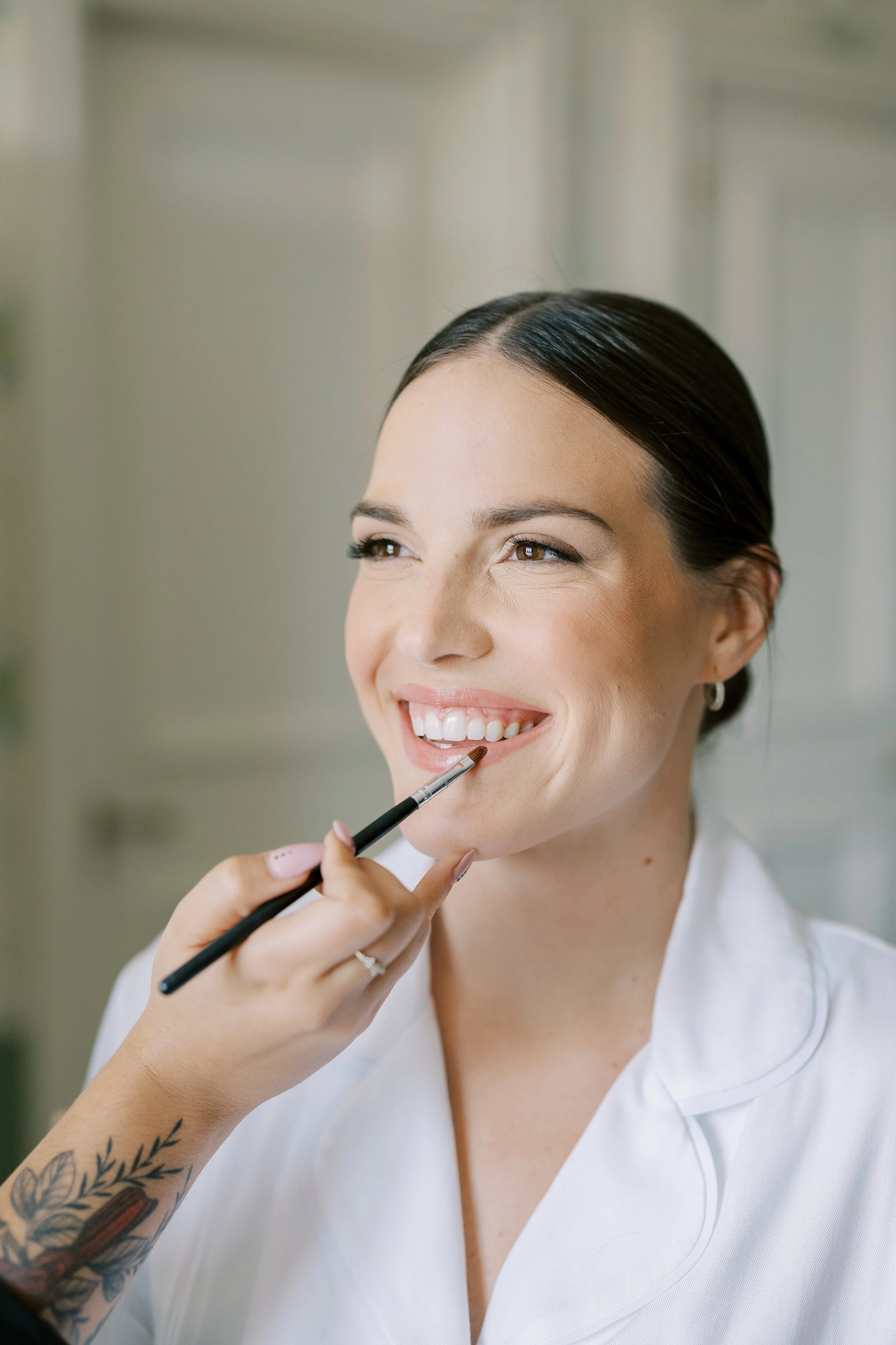 A woman smiling as a makeup artist applies lip gloss with a brush, in a bright room.