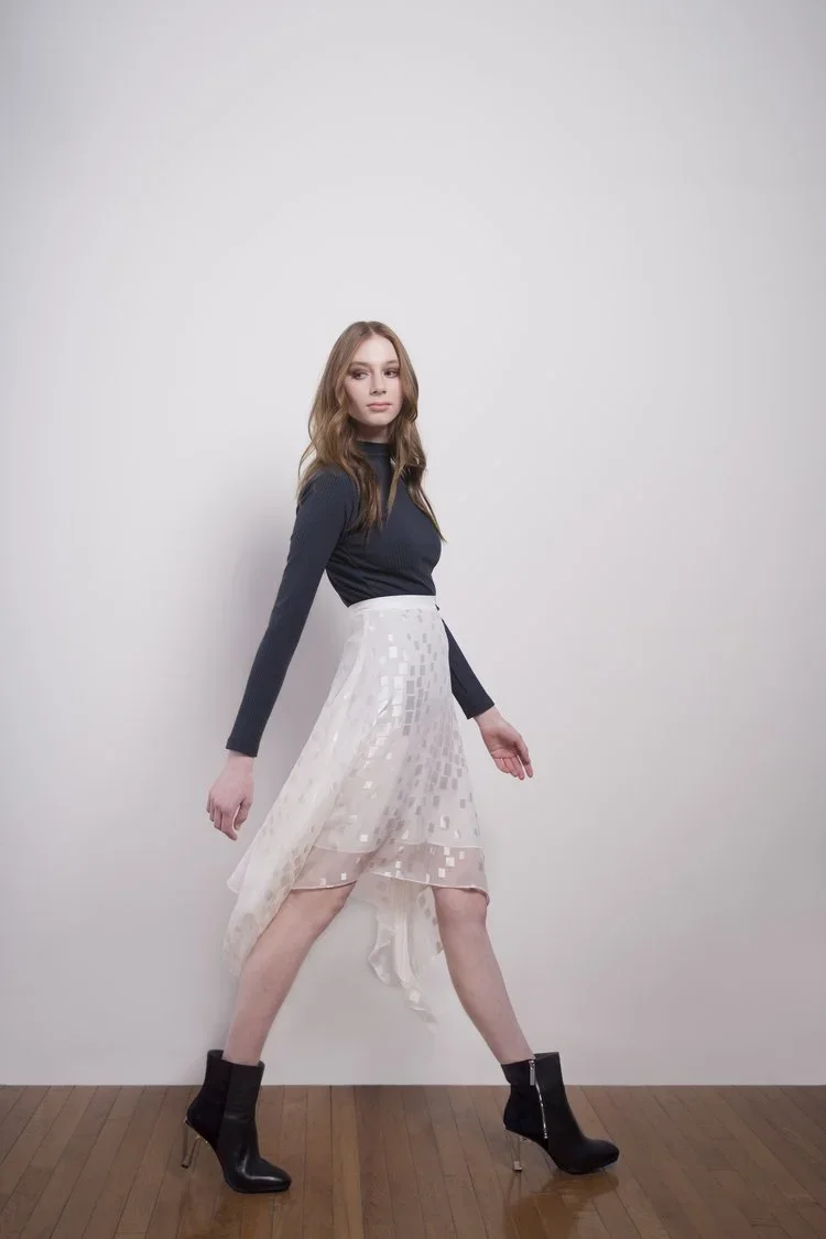A young woman with long wavy brown hair standing against a plain white background. She is wearing a navy blue long-sleeve top, a white semi-sheer skirt with a checkered pattern, and black high-heeled ankle boots. She poses with her legs apart and her hands relaxed at her sides.