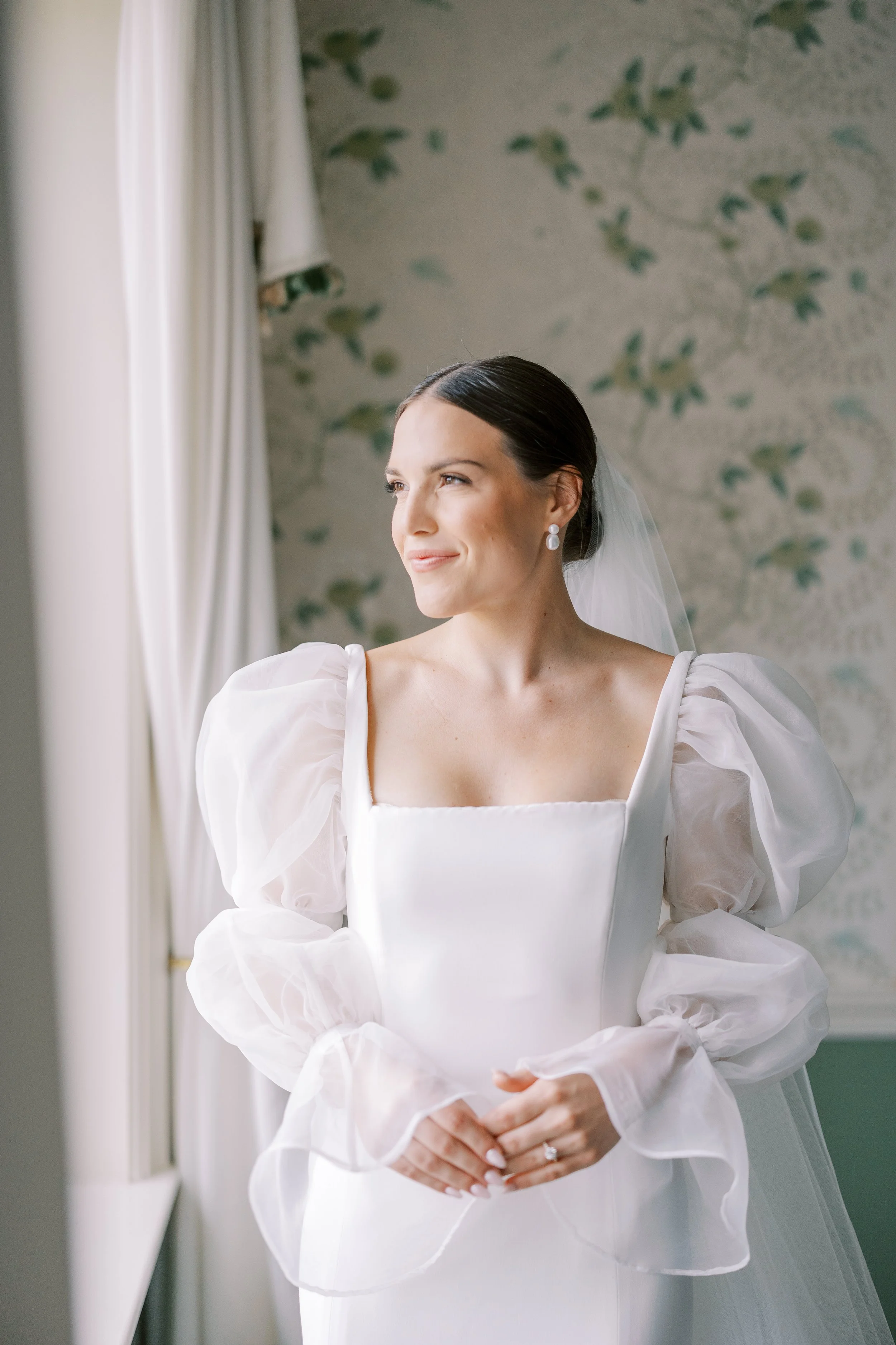 A bride in a white wedding dress with puffed sheer sleeves, standing near a window and looking outside with a gentle smile.