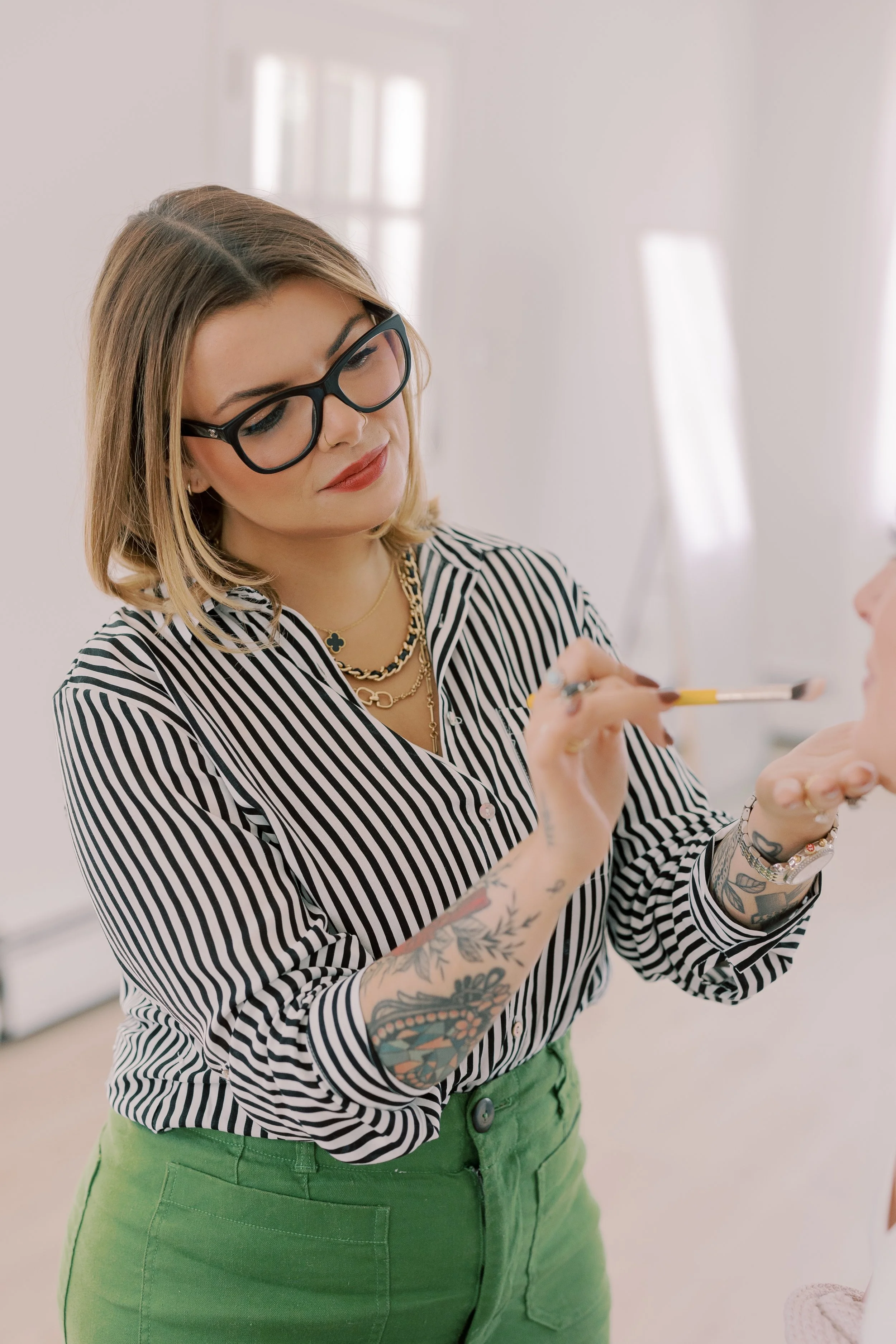 A woman with short blond hair, wearing glasses, a black and white vertically striped blouse, and green pants, applies makeup with a brush on another person's face in a brightly lit room.