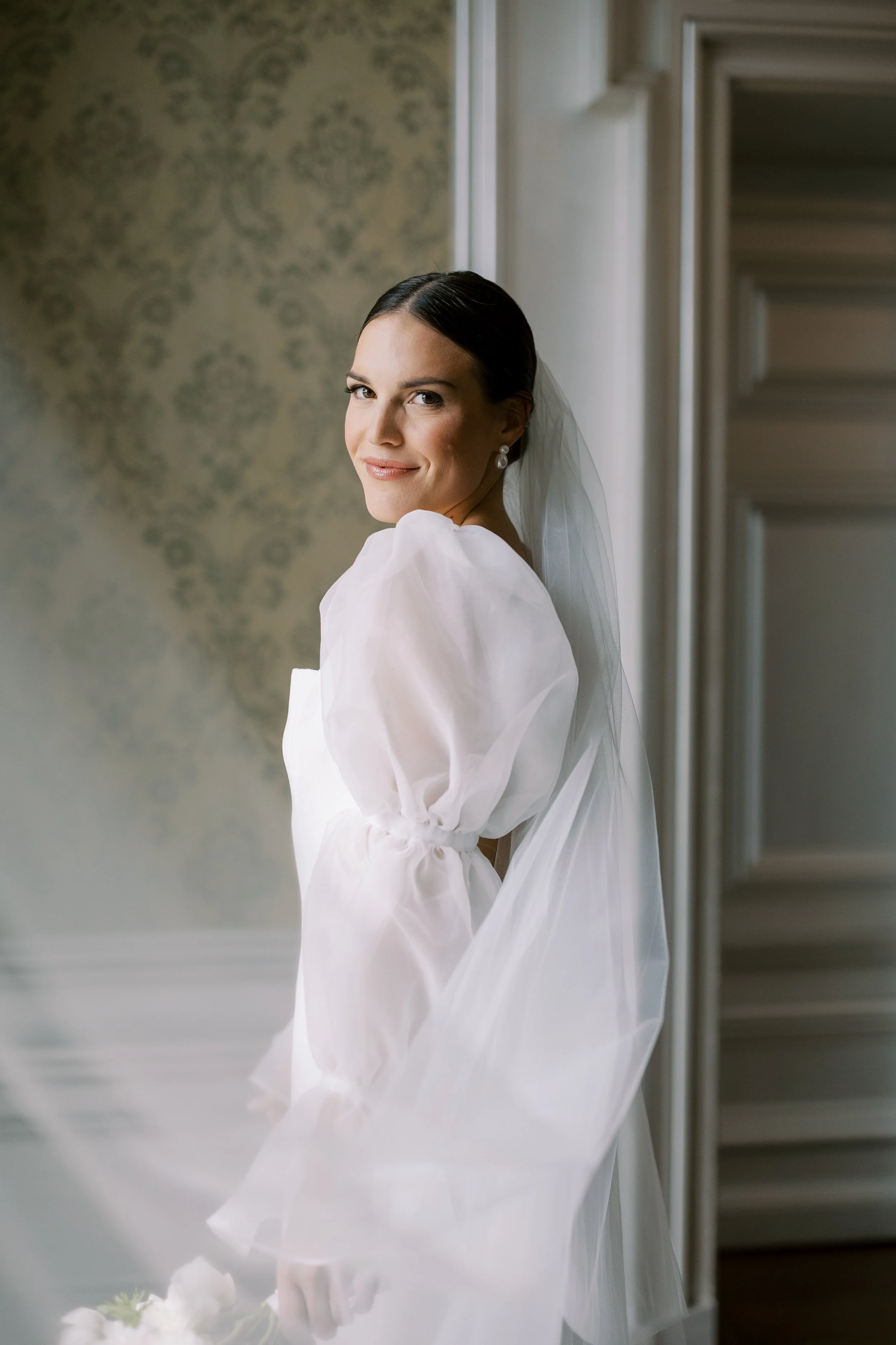 Bride in a white gown with puffed sleeves, smiling in a beautifully decorated room.