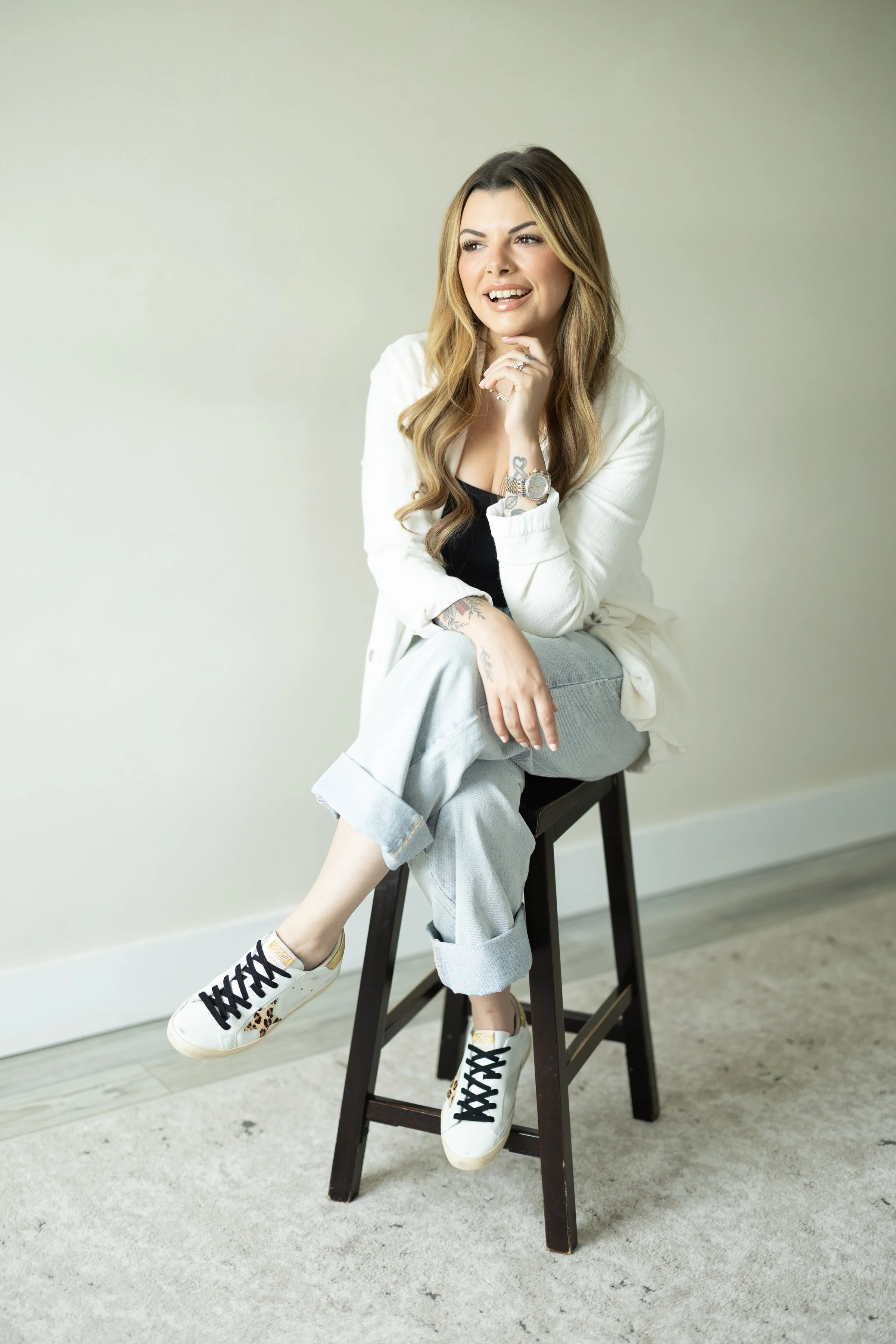 A young woman with long wavy hair sitting on a black wooden stool, wearing a white jacket, black top, light blue jeans, and white sneakers with leopard print accents, posing with a playful smile and hand on chin, against a plain light-colored background.