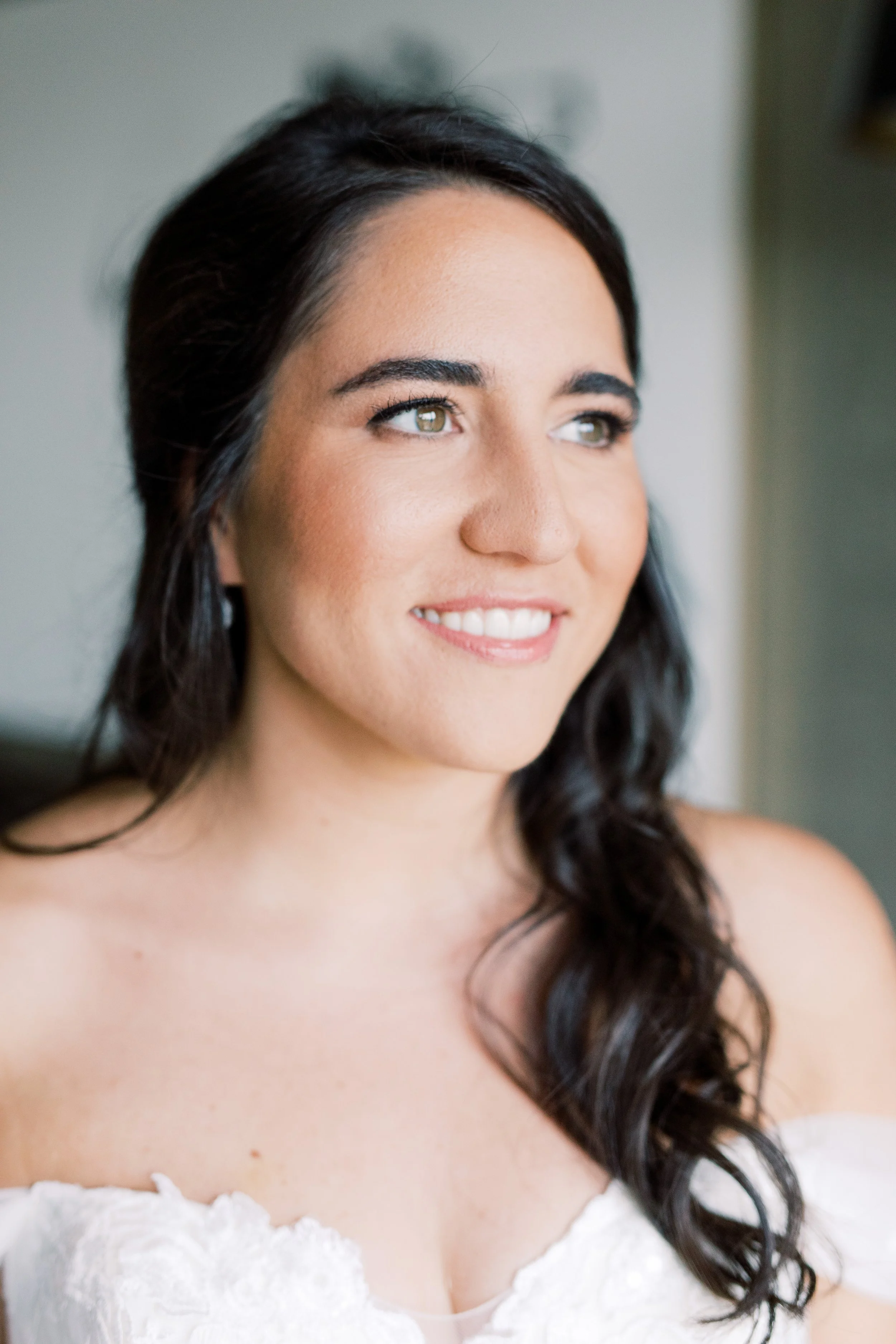 A woman with dark hair and brown eyes smiling, wearing a white dress, indoors with a neutral background.