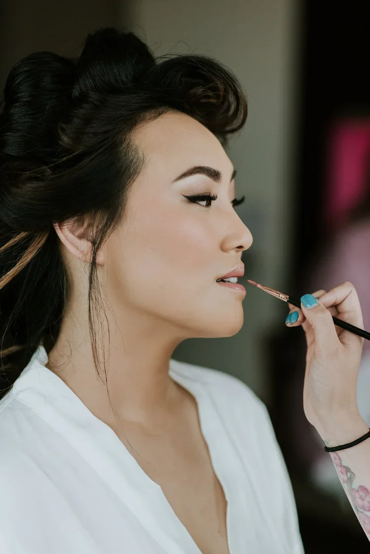 Close-up of a woman with styled hair and makeup getting her lips painted with a makeup brush.