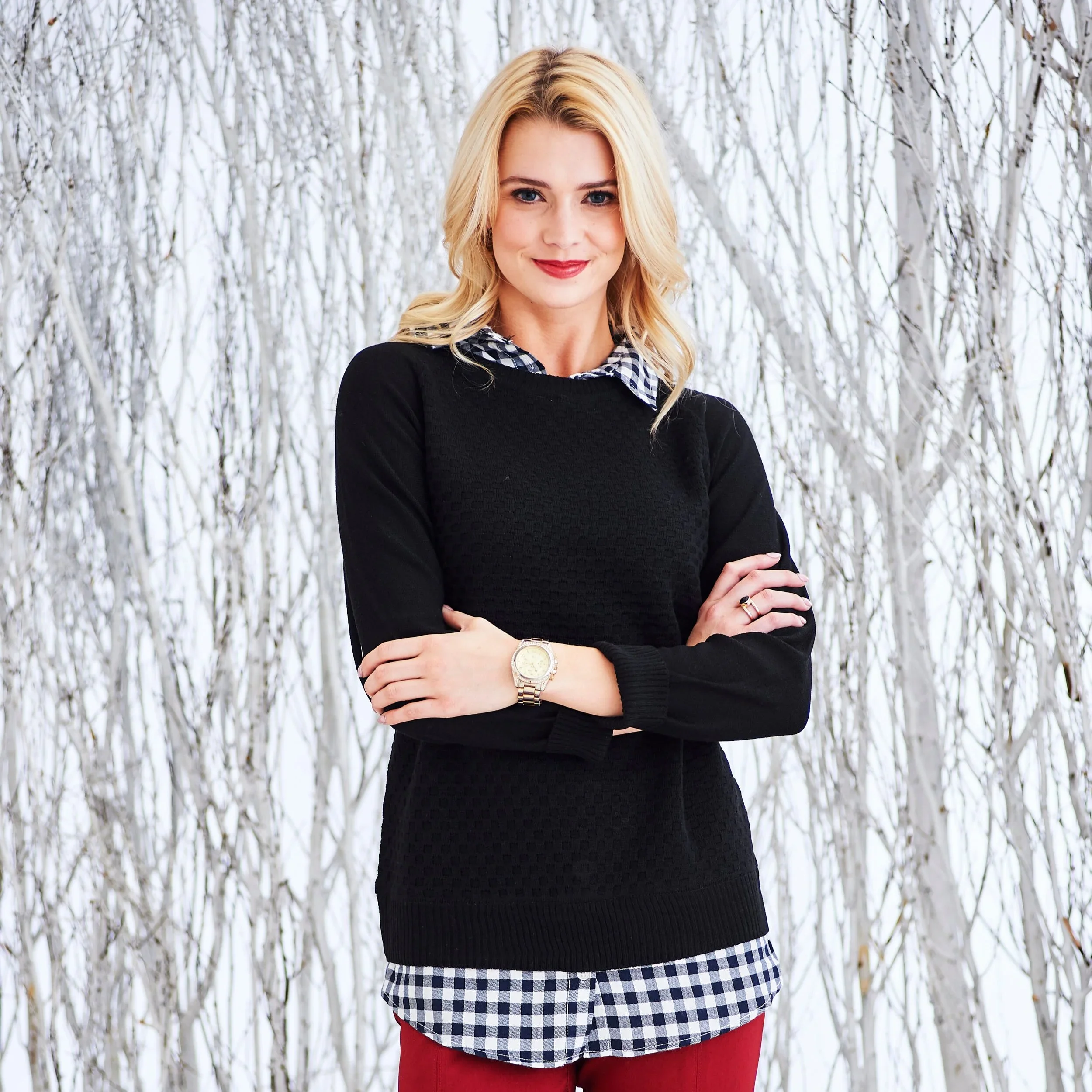 A woman with blonde hair smiling and standing with arms crossed in front of a snowy forest background.