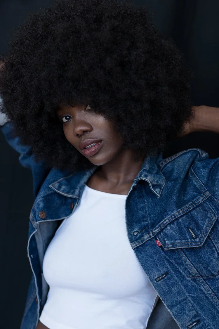 Close-up of a woman with a large afro hairstyle wearing a denim jacket and white top, posing against a dark background.