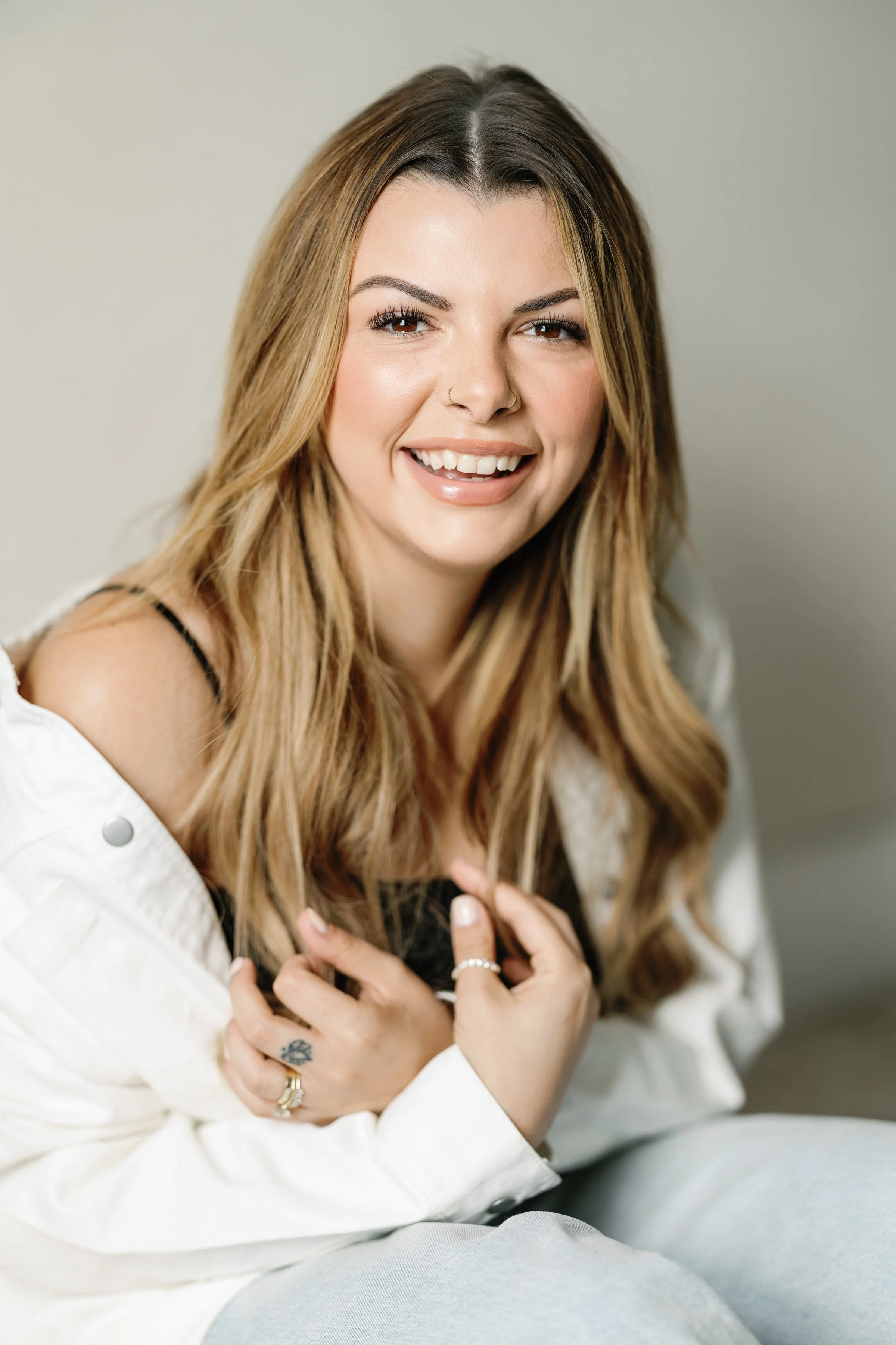 A woman with long, wavy, blonde hair smiling and looking at the camera. She is wearing a white jacket off her shoulders and has rings on her fingers.