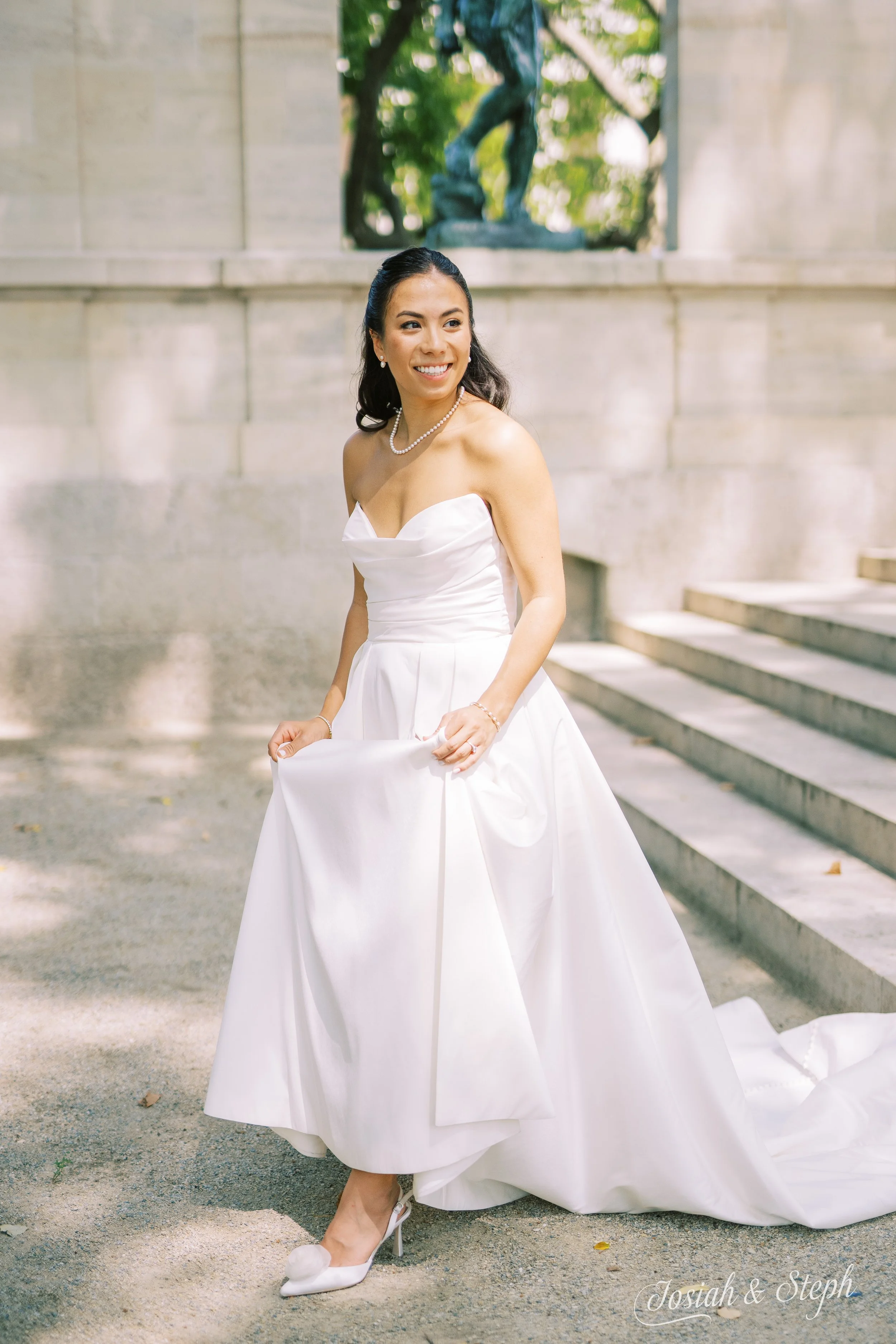 A smiling woman in a white wedding gown and heels outdoors, holding her dress in front of a stone wall with stairs and a bronze statue in the background.