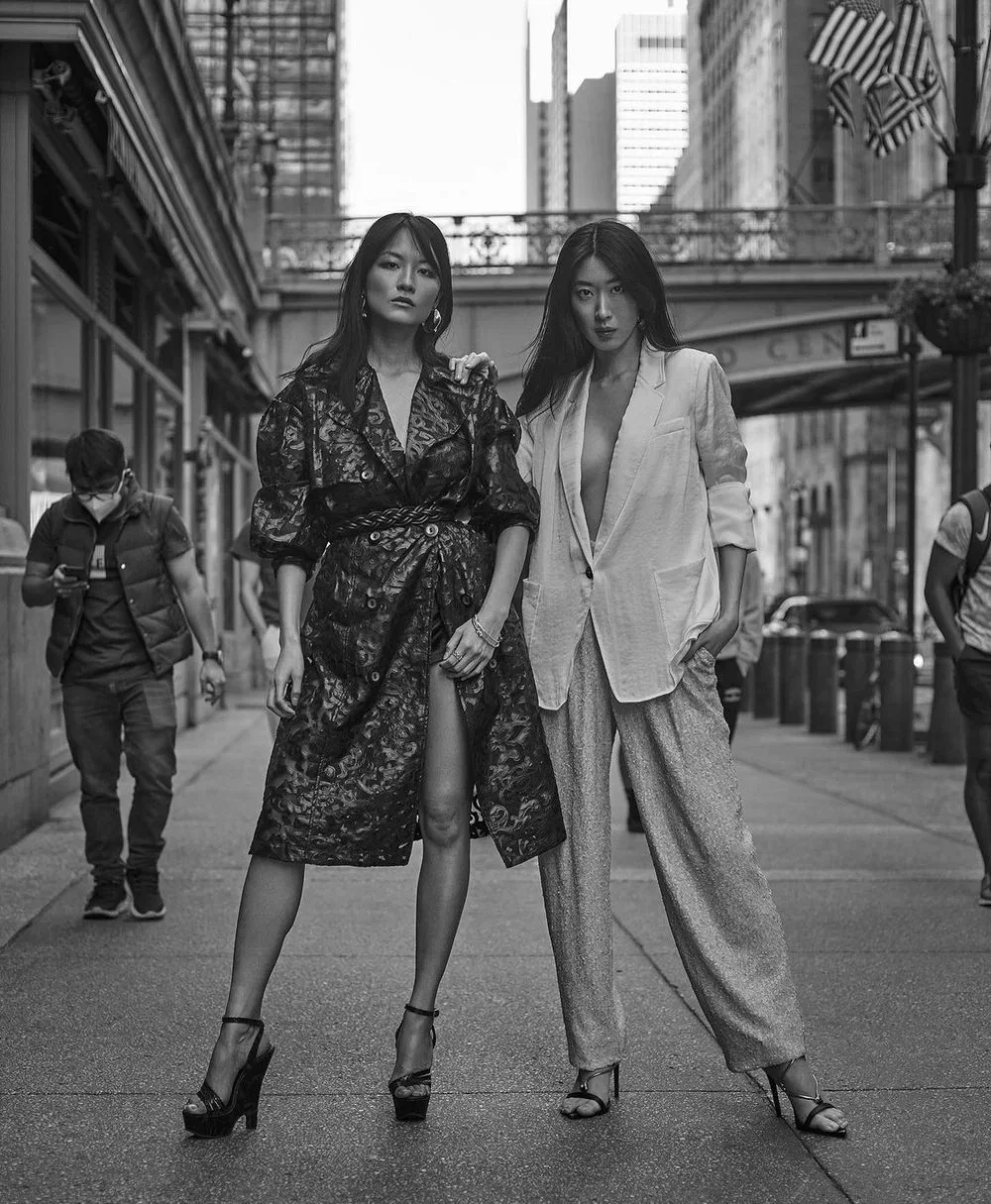 Two women posing confidently on a city street, dressed in fashionable high-end outfits, with skyscrapers and pedestrians in the background.