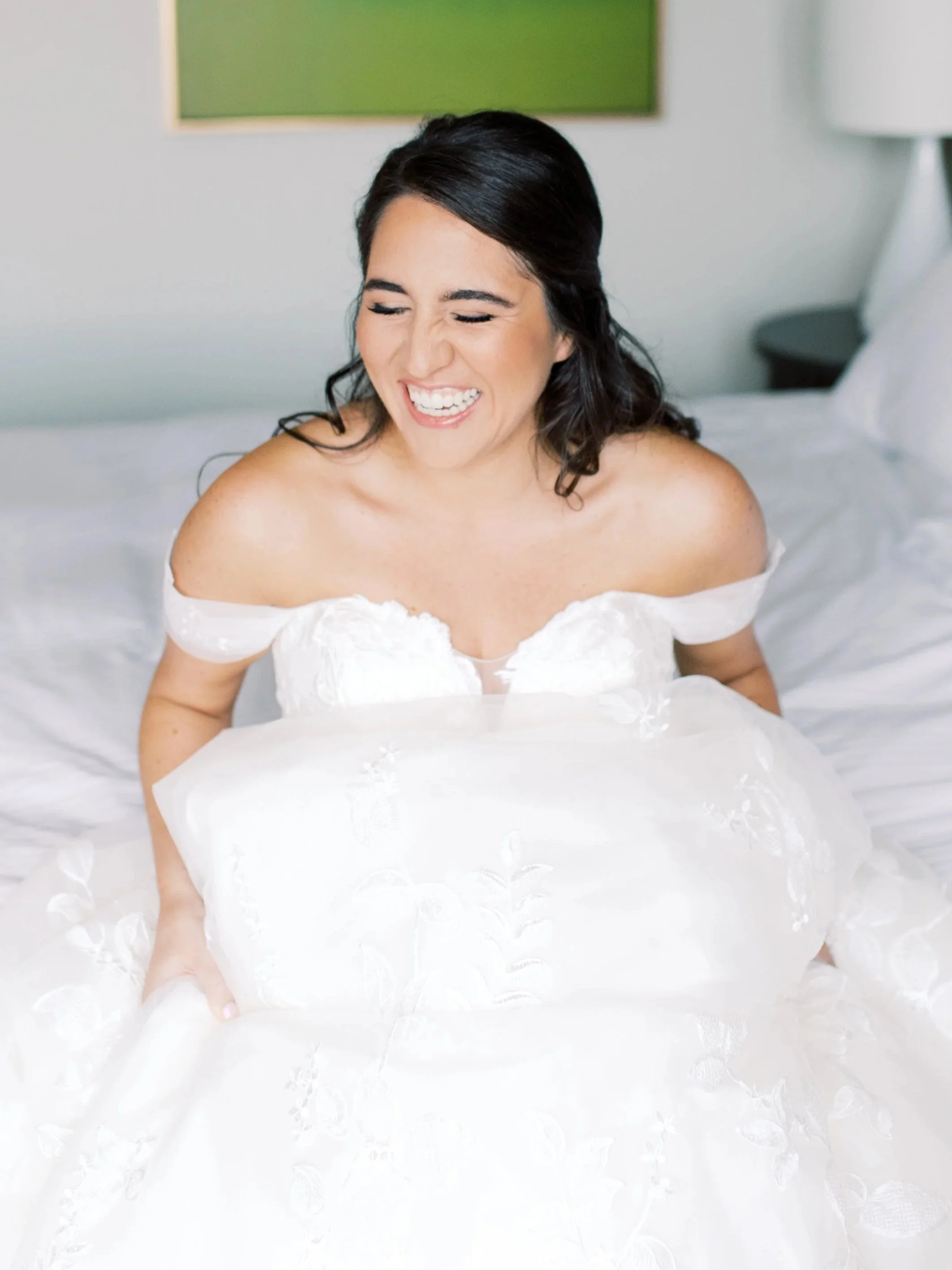 A woman in a white off-shoulder wedding dress sitting on a bed, smiling with eyes closed.
