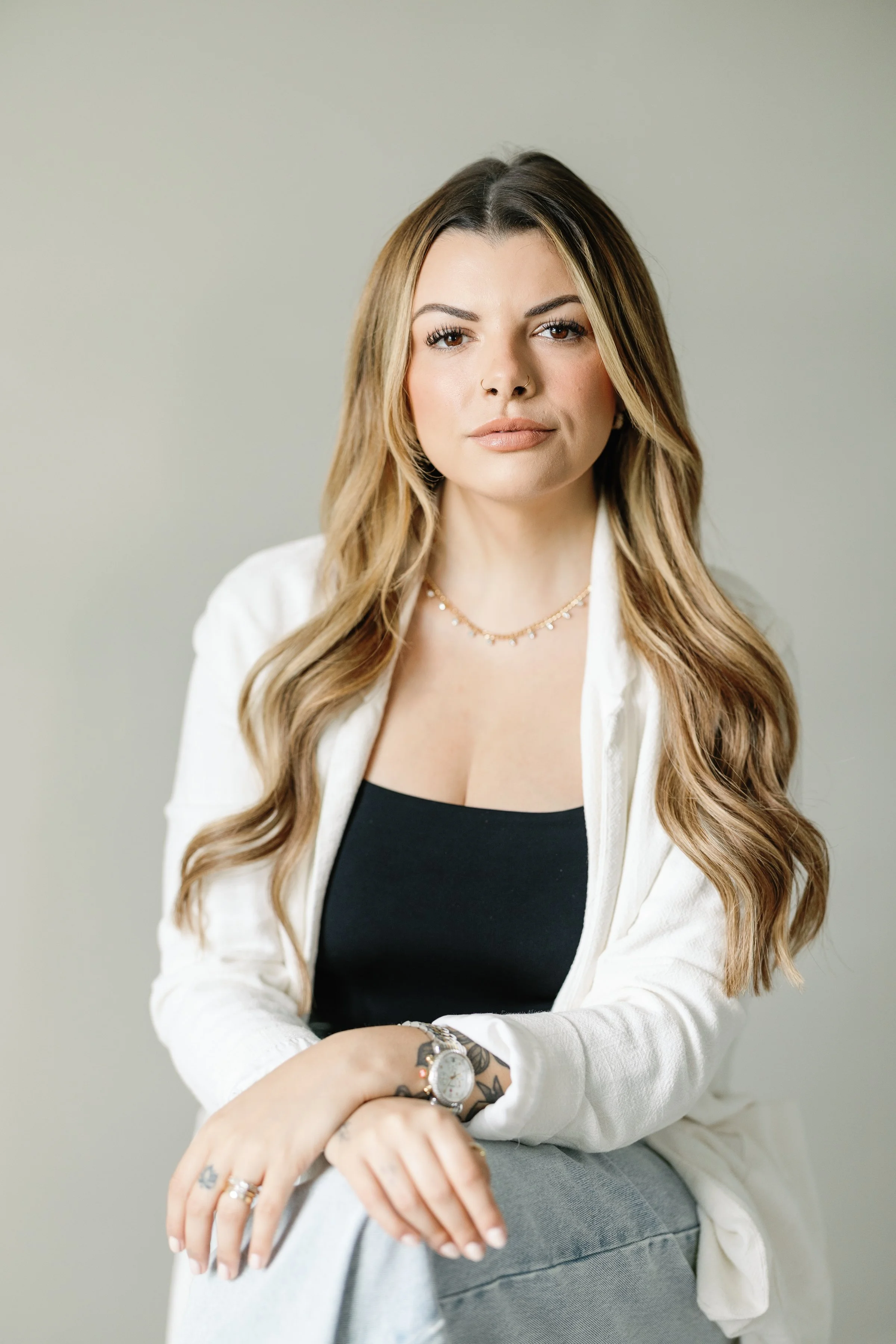 A woman with long, wavy blonde hair and light skin, wearing a black top, white blazer, gray pants, a watch, rings, and a delicate necklace, sitting against a plain gray background.