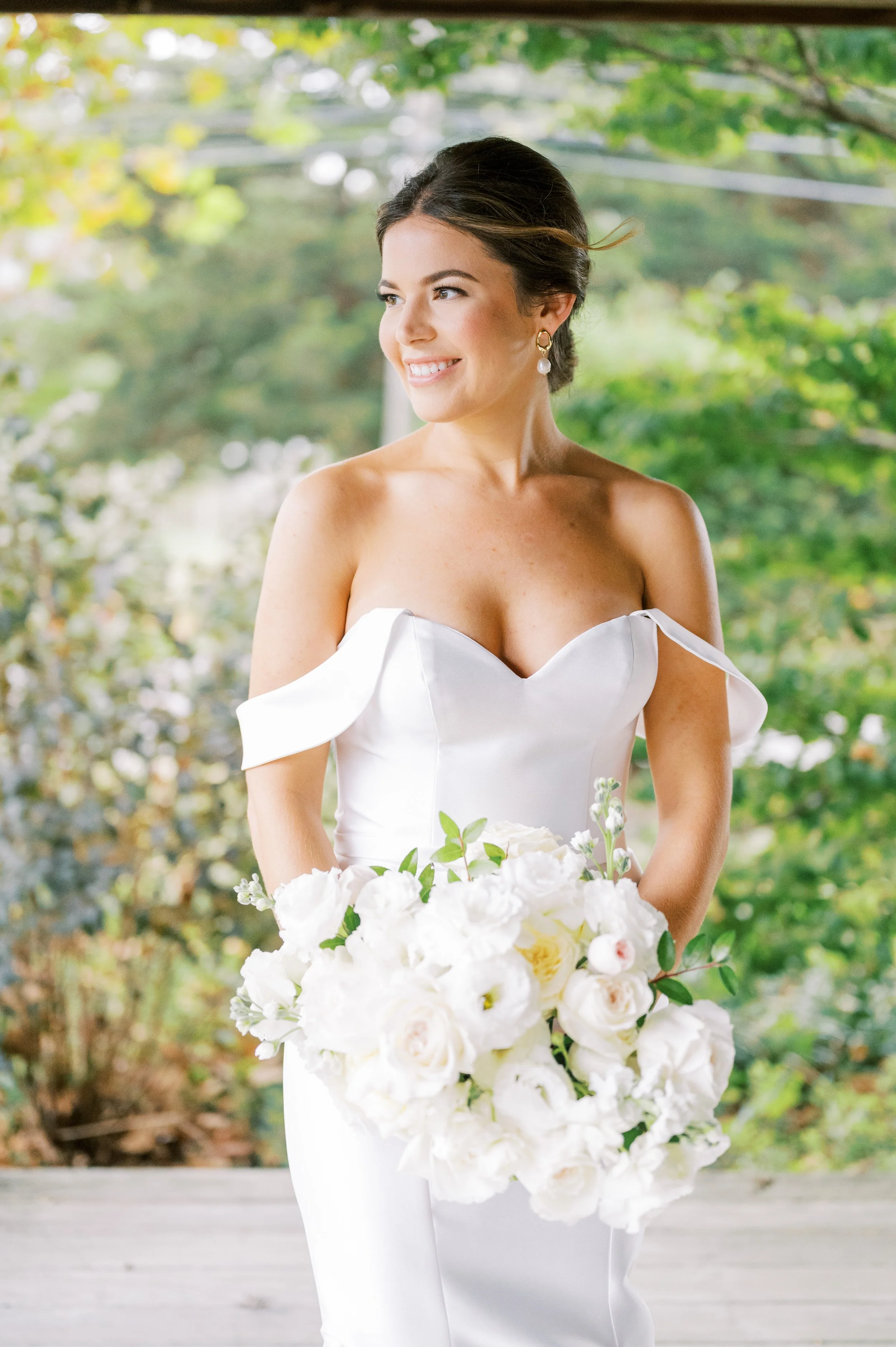 Bride in white off-shoulder gown holding a large bouquet of white flowers, standing outdoors with green foliage background.