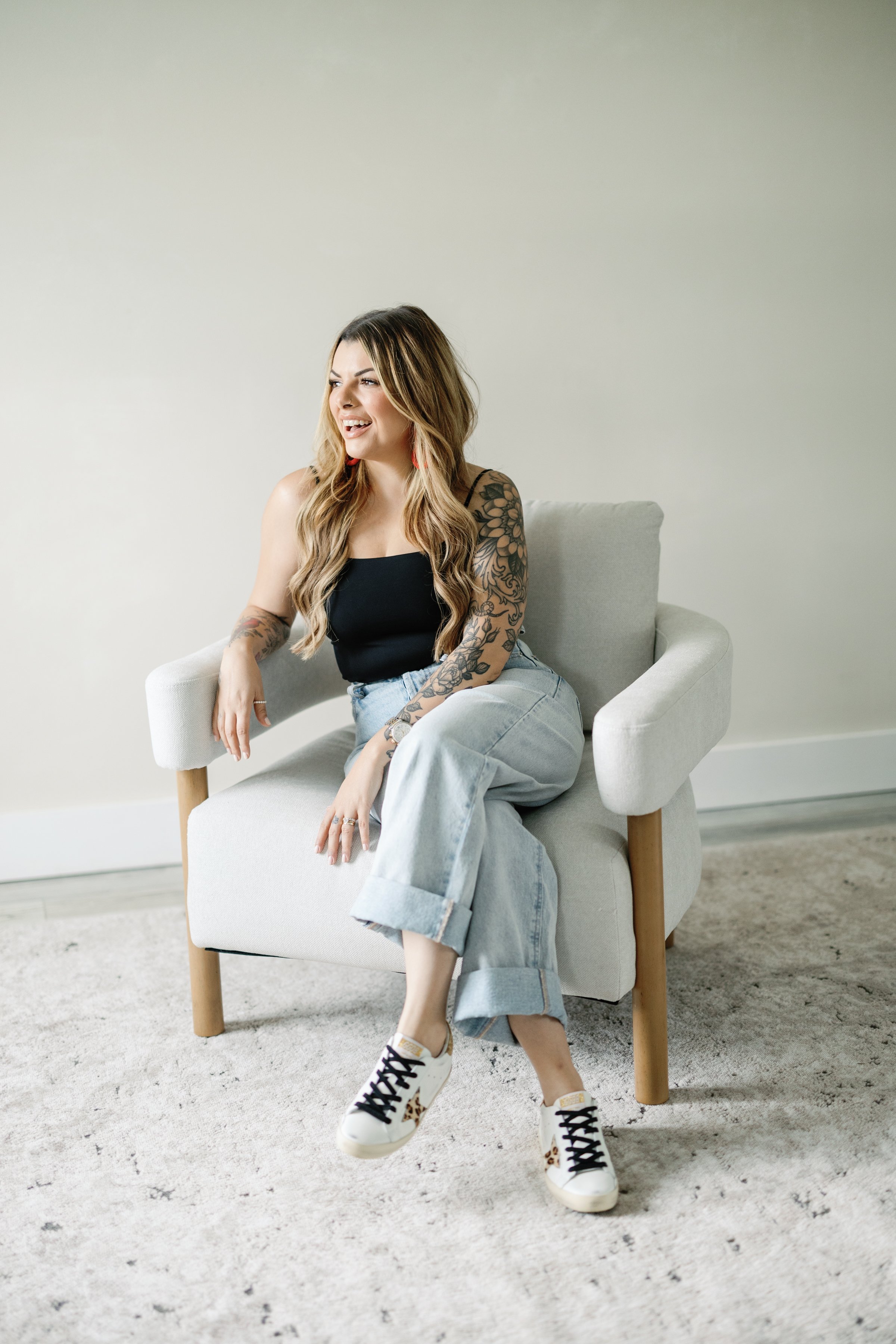 A woman with long, wavy hair and tattoos on her arms, sitting on a light-colored armchair in a minimalistic room. She is smiling, wearing a black tank top, light blue jeans, and white sneakers with leopard print detail.