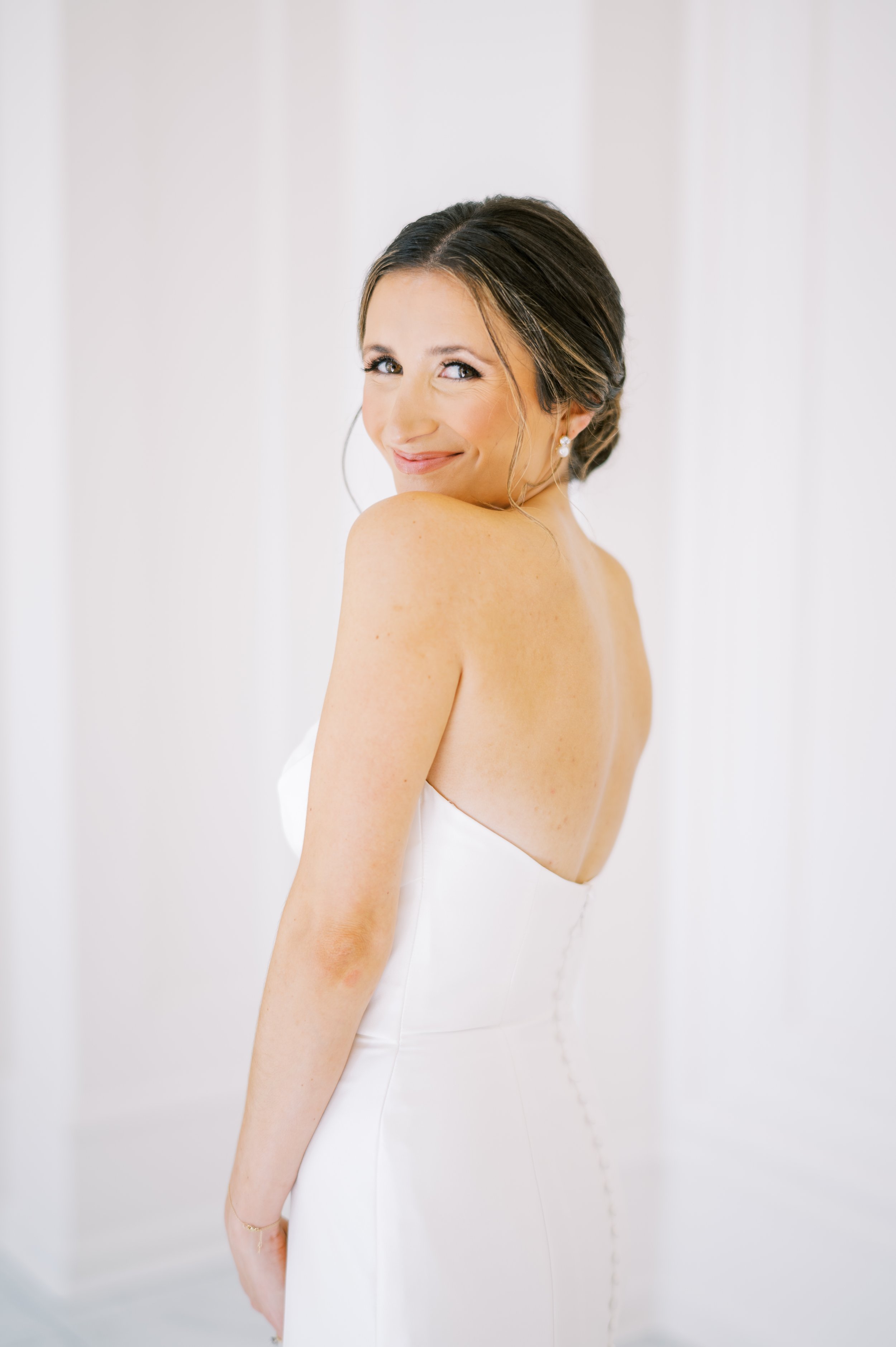 A smiling woman in a white strapless wedding gown with a low back, standing in a bright, white room.