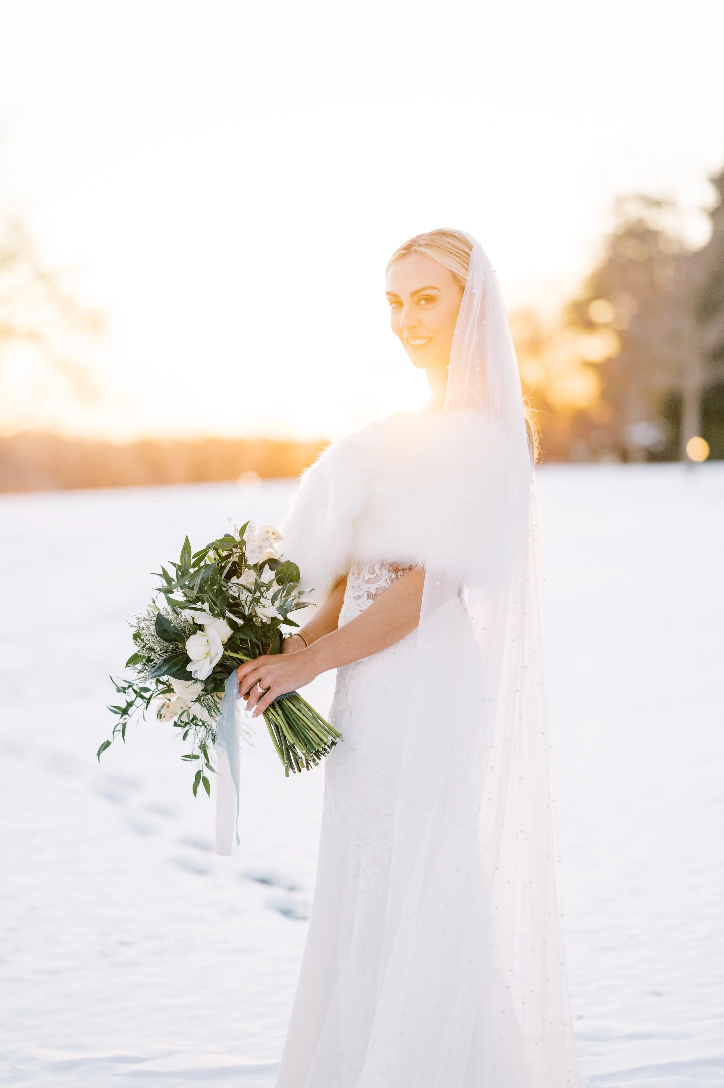 Bride in a white wedding dress and veil holding a bouquet of white flowers in a snow-covered field during sunset.