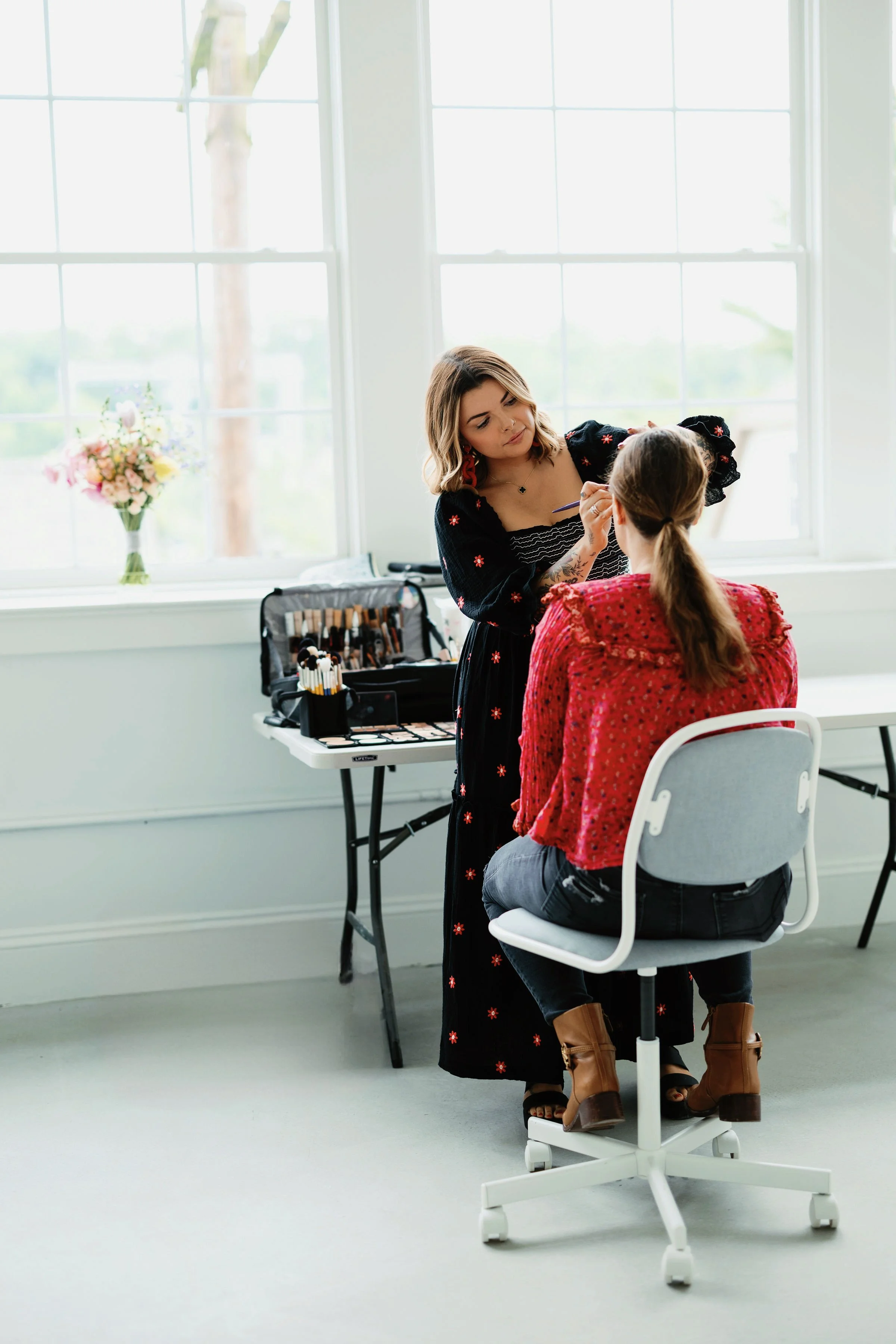 A woman is sitting on a white chair while a makeup artist applies makeup to her face. The makeup artist is standing next to her, with a makeup case open on a table. There is a window behind them with a vase of flowers on the windowsill.