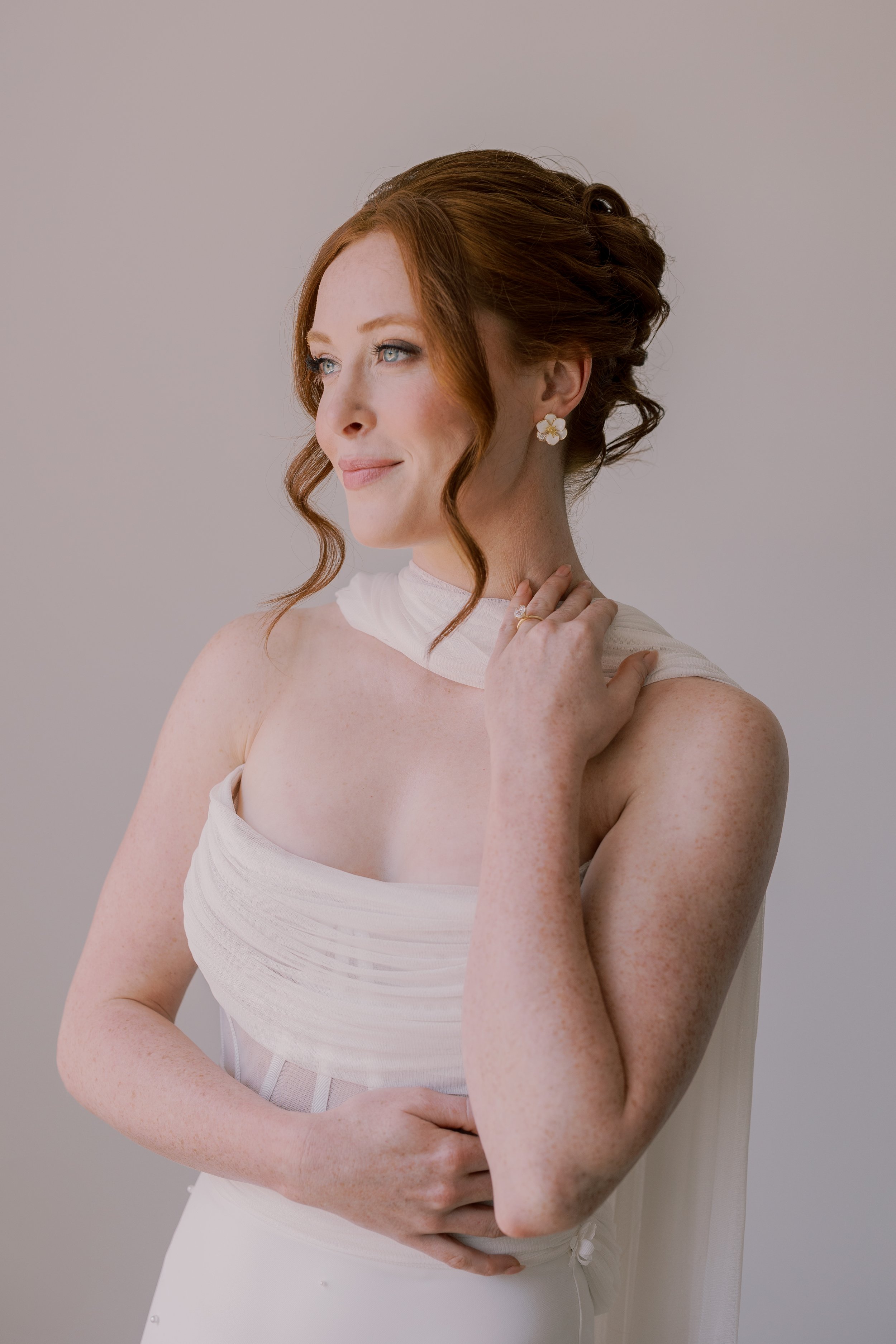 A bride with red hair styled in loose curls, wearing a white strapless wedding gown, with floral earrings, and a gentle smile, standing against a plain background.